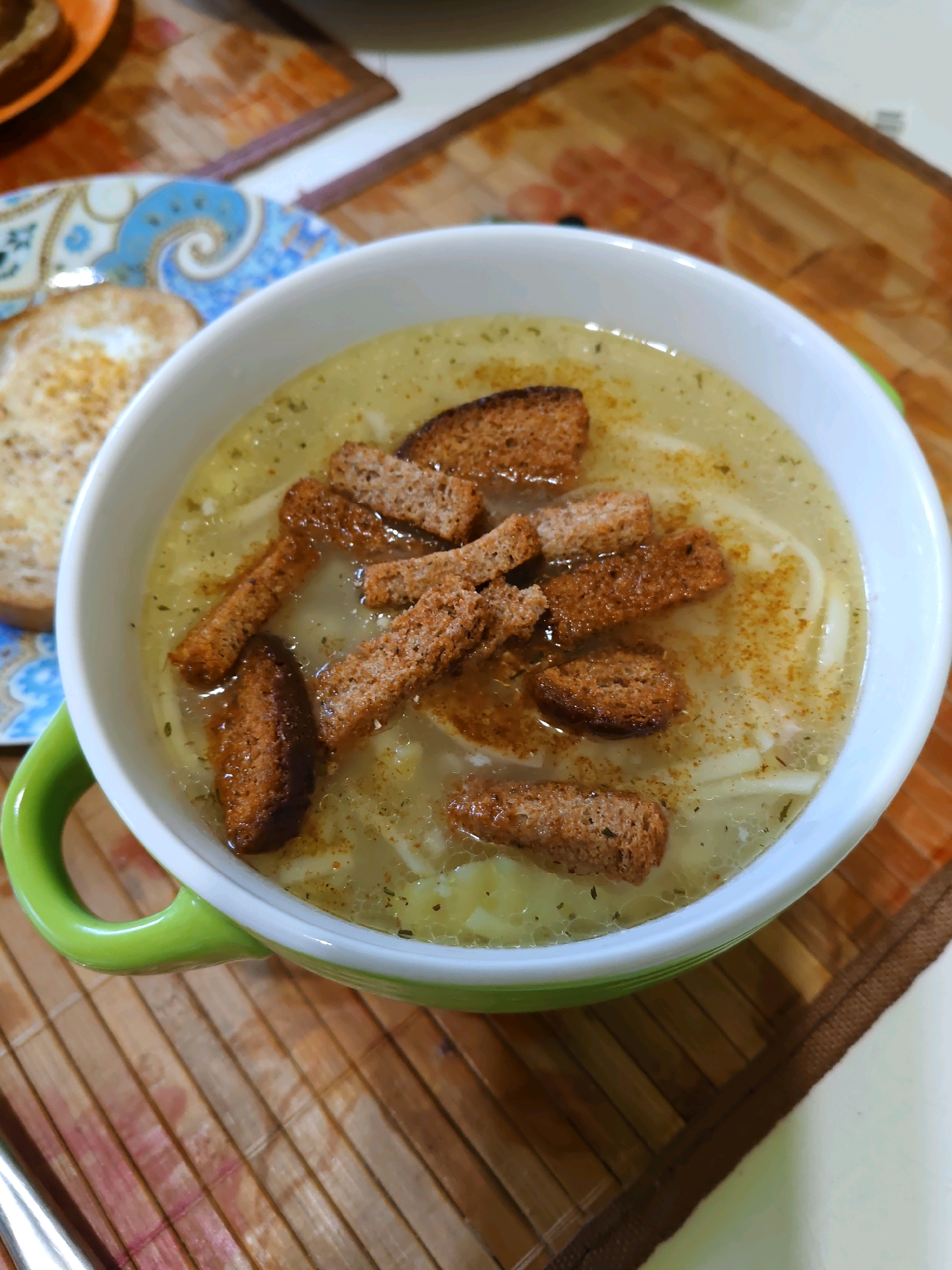 A close-up, overhead shot shows a bowl of soup topped with croutons on a wooden placemat. The soup appears to be a light-colored broth with noodles and possibly some diced vegetables. The croutons are small, rectangular, and toasted brown. To the left of the soup bowl, a plate with two slices of toasted bread is visible. The placemat is made of light-colored wood with a visible grain. The background is blurred, but appears to be a table setting. The image was taken in Rostov-on-Don, Russia.