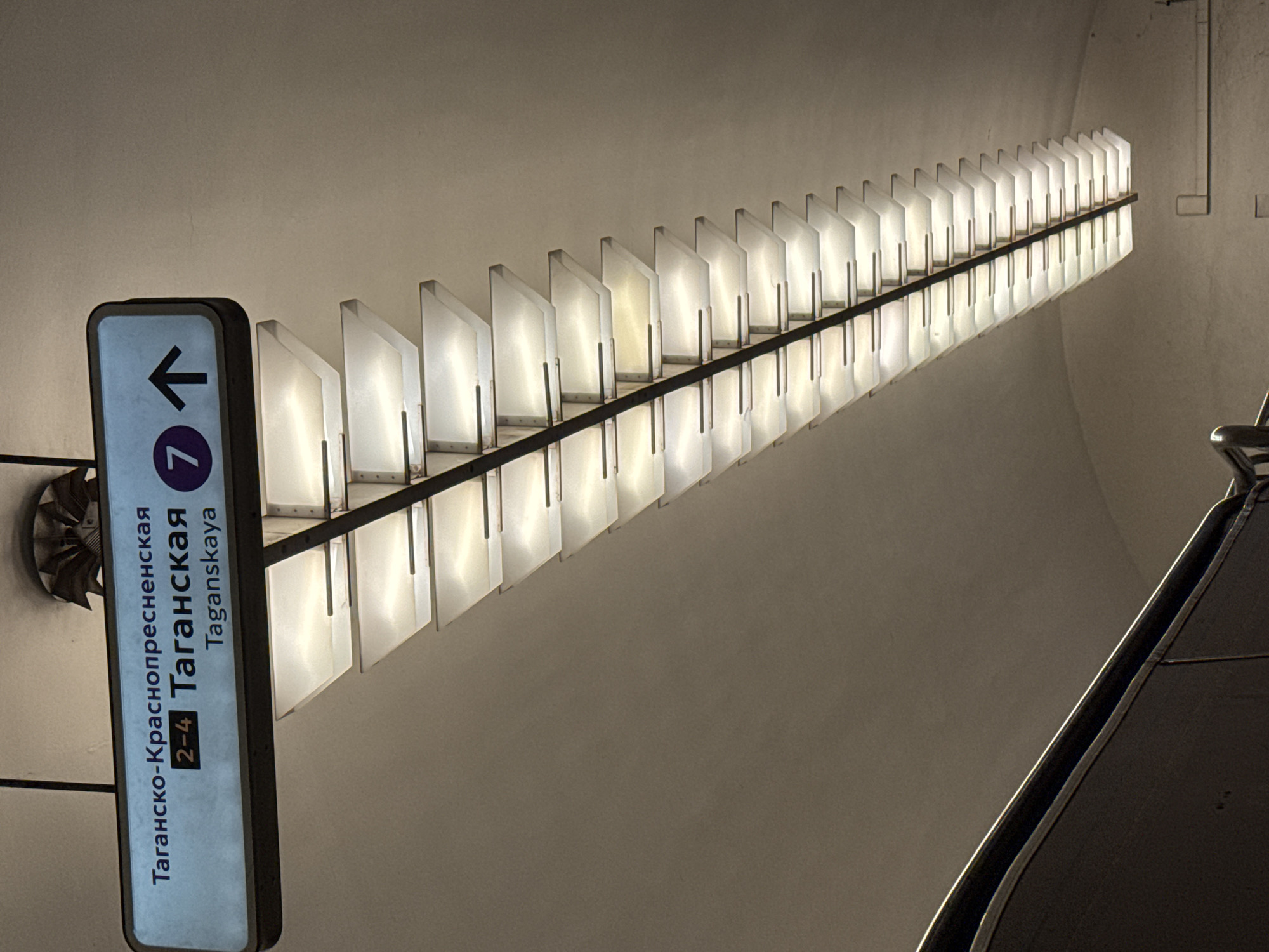 The image shows a ceiling sign in a subway station. The sign is rectangular and illuminated, with blue text. The text is in Russian and English. The Russian text reads "Таганско-Краснопресненская 2-4 Таганская Taganskaya". To the right of the text, there is a purple circle with the number "7" inside it, followed by a black arrow pointing upwards. Below the sign, a long, tiered light fixture extends horizontally. The fixture is made of metal and has multiple rectangular, translucent panels that emit a warm light. The ceiling appears to be arched and made of a light-colored material. In the lower left corner, a dark-colored handrail is partially visible, suggesting a staircase or escalator is nearby. The overall environment is consistent with a modern subway station.