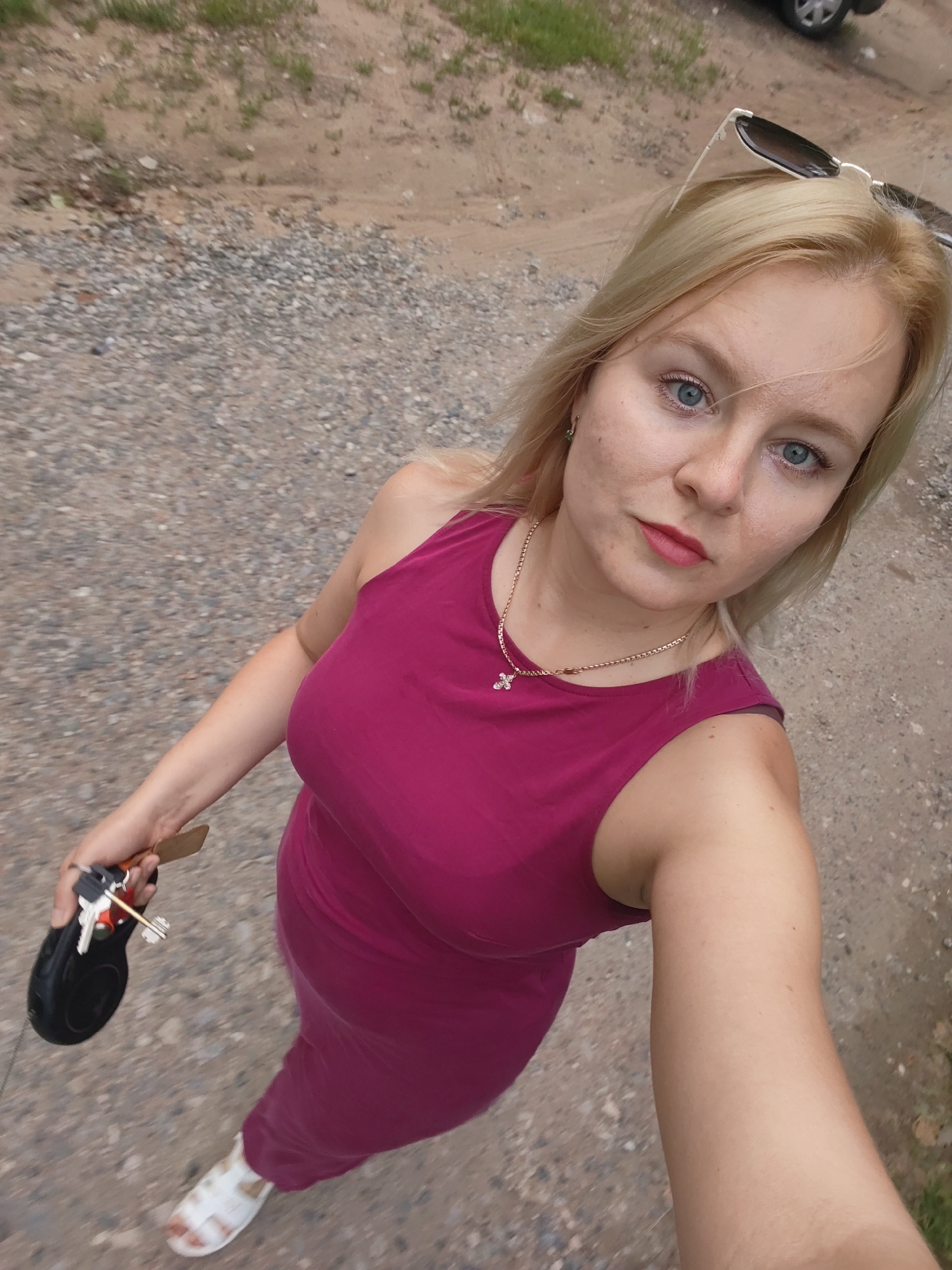 A woman with blonde hair and blue eyes, wearing a pink dress and white sandals, stands on a gravel path. She is holding keys and a retractable leash in her right hand. Sunglasses are perched on her head. The background shows a dirt and grass embankment, and a car is visible in the upper right corner. The scene appears to be an outdoor location in Izhevsk, Russia.