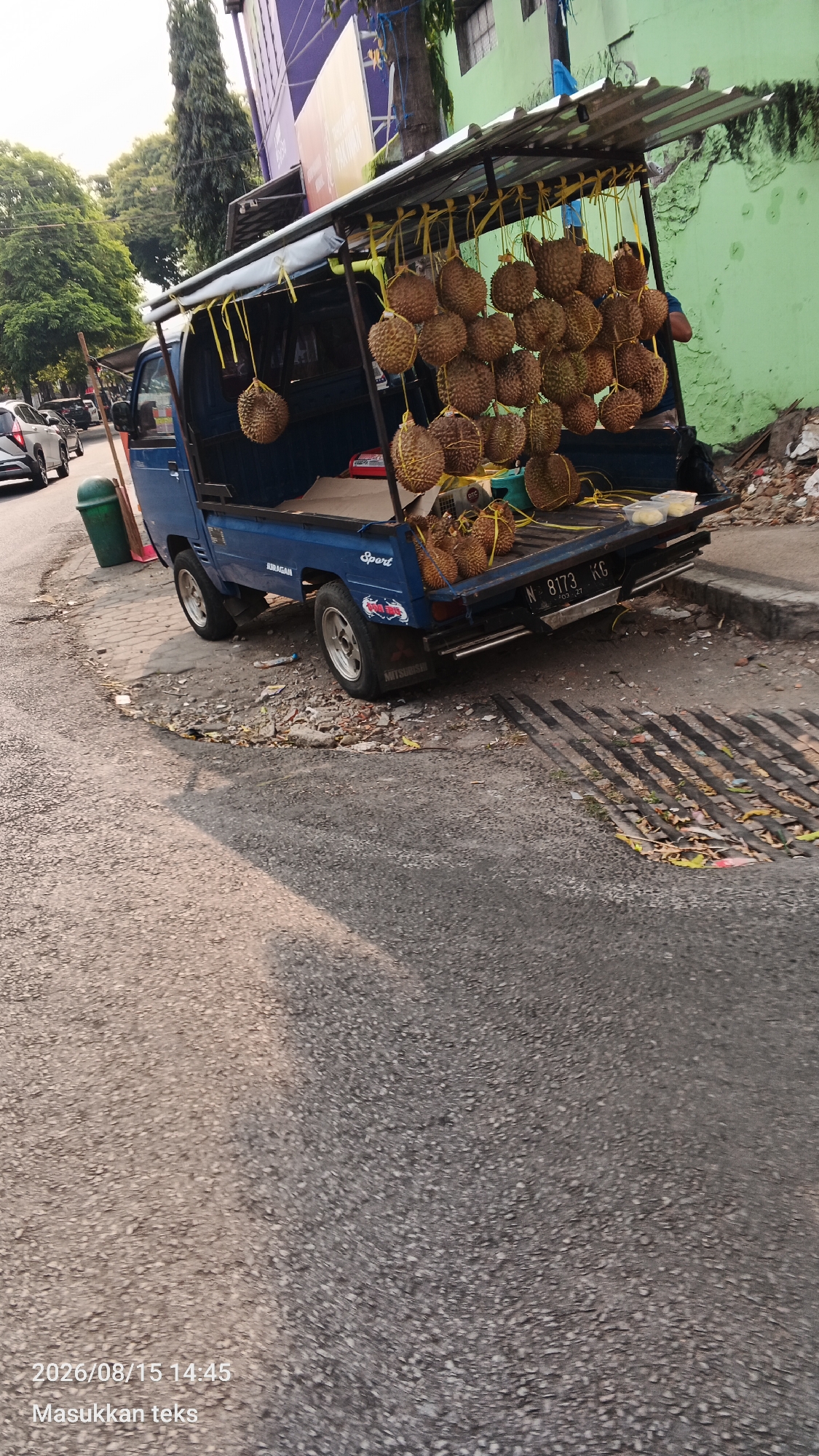 An outdoor, eye-level shot captures a blue utility truck parked on the side of a street, likely in Malang, Indonesia, given the license plate. The truck bed is laden with durian fruits, some of which are hanging from a makeshift canopy above. The fruits are spiky and brown, characteristic of durian. A person, partially visible, stands near the truck. The surrounding environment includes a green wall, trees, and the asphalt road. The ground is littered with debris, and a metal grate is visible on the road. The image also displays a timestamp in the lower left corner: "2026/08/15 14:45" and the text "Masukkan teks".