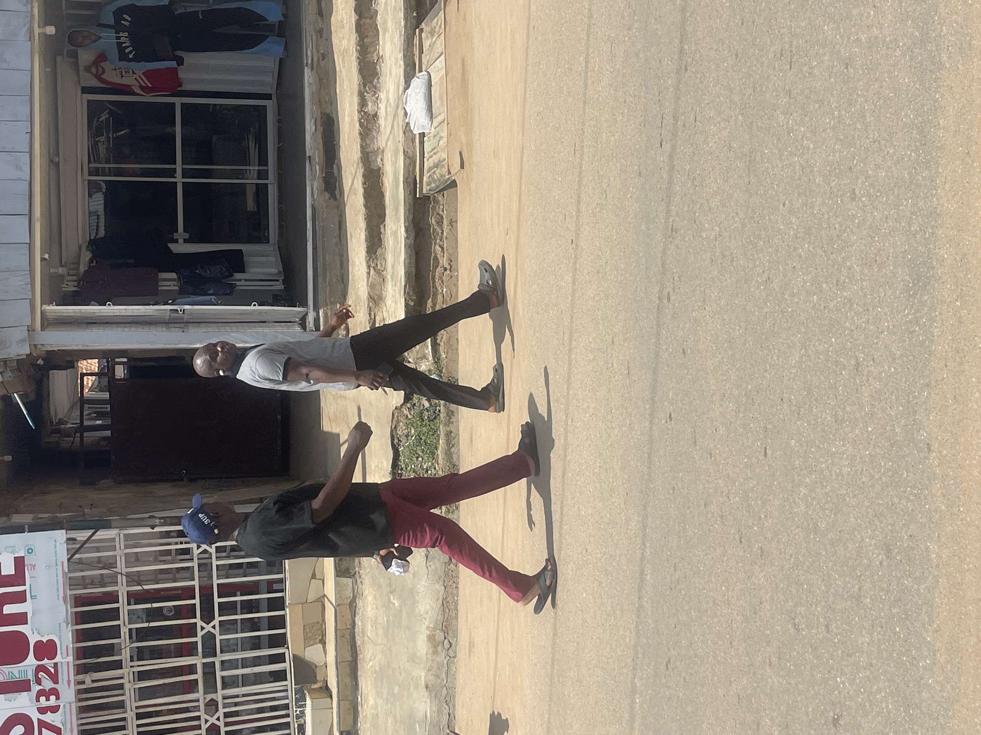 The image shows two men walking on a street in Abuja, Nigeria. The man in the foreground is wearing a dark t-shirt, burgundy pants, and a blue baseball cap. He is walking with his left leg forward. The man behind him is wearing a grey t-shirt and dark pants, and is walking with his right leg forward. Both men are walking on a paved street. To the left of the street is a building with a shopfront. The shop appears to be selling clothing, with items displayed in the window and hanging outside. There is a sign above the shop entrance with red and white lettering. The building is made of concrete and has a rough texture. The sun is shining, casting shadows on the ground, suggesting it is daytime. The weather appears to be warm and sunny. The overall scene depicts a common street activity in an urban environment.