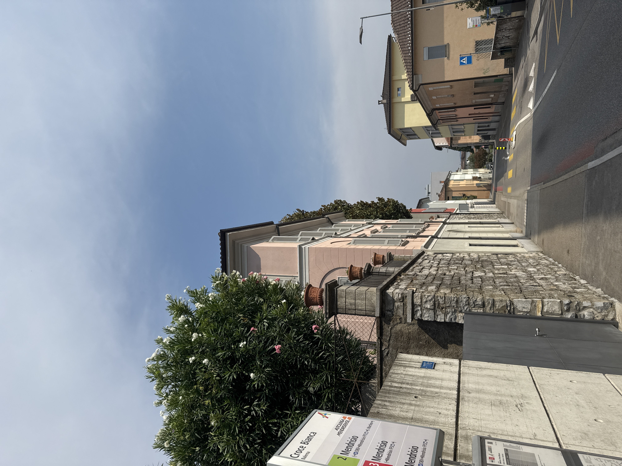 The image shows a street scene in Balerna, Switzerland. On the left, a bus stop sign displays "Croce Bianca" and lists destinations, including "Mendrisio." A large tree with white flowers is in the foreground, to the right of the bus stop. Behind the tree is a pink building with windows. A stone wall runs along the sidewalk, and a gray metal gate is visible. The street is paved, with yellow and white markings. Several buildings line the street, varying in color and architectural style. The sky is blue with some clouds.
