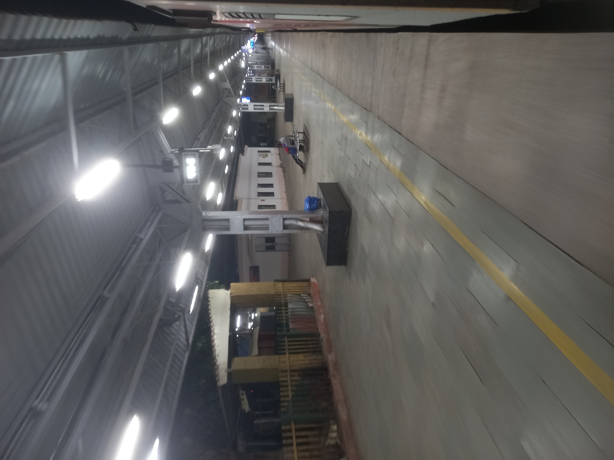 The image depicts an empty train platform in Igatpuri, India, at night. Overhead lights illuminate the platform, which is long and narrow. A yellow safety line runs along the edge of the platform. There are waiting areas with benches. The platform is under a metal roof. The perspective suggests the photo was taken from a train car.