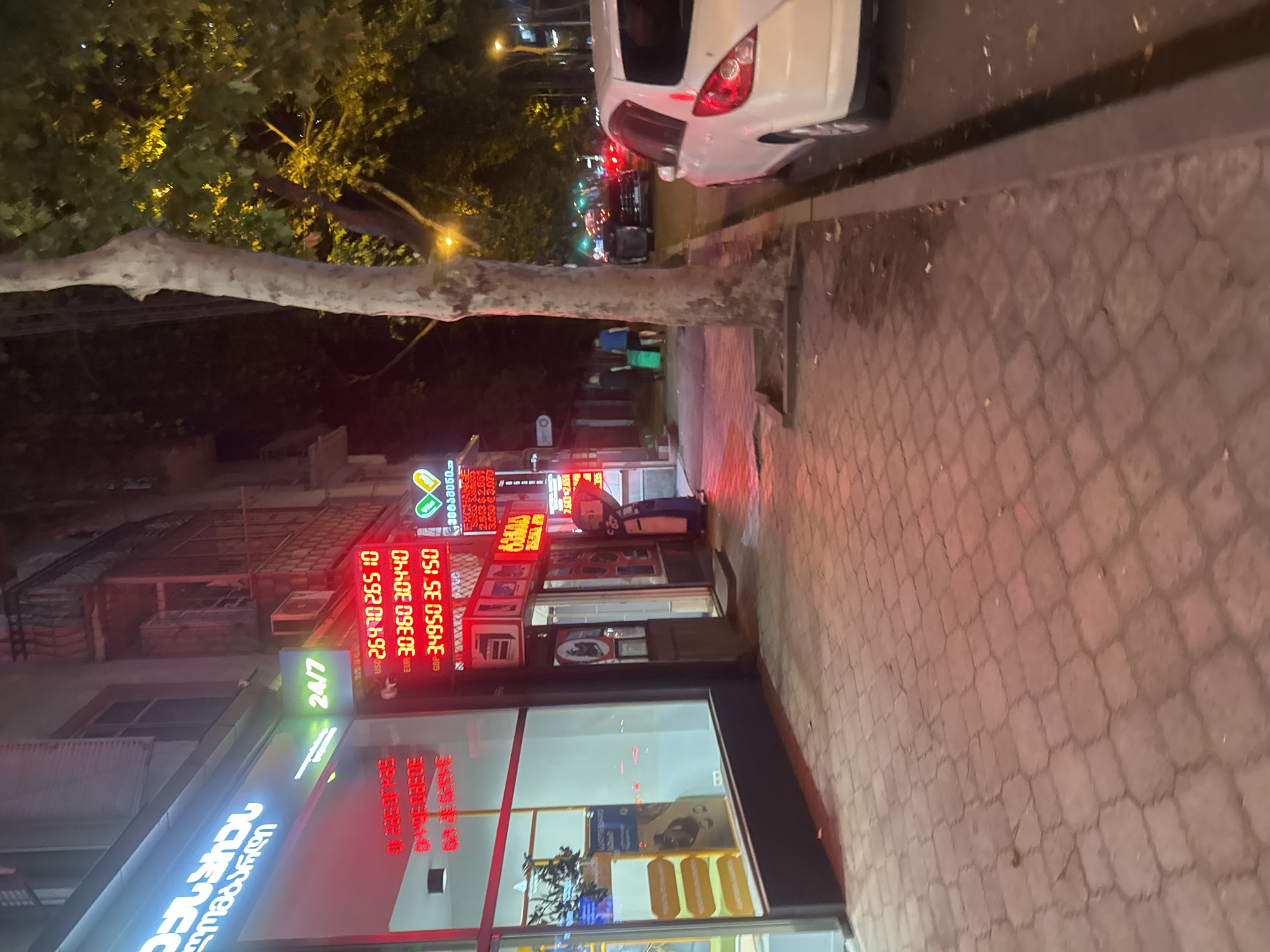 The image depicts a street scene in Tbilisi, Georgia, at night. A sidewalk paved with hexagonal bricks runs along the left side of the frame, illuminated by the red neon glow of a currency exchange office. The office displays exchange rates for USD, EUR, and GBP, indicating "24/7" operation. To the right of the exchange office, a tall tree with a pale trunk stands beside the sidewalk. Beyond the tree, a white car is parked on the street, with other vehicles and traffic lights visible in the background, suggesting a busy urban environment. The building facade on the left features windows with displays and signage in Georgian script. A person can be seen walking on the sidewalk in the mid-ground, pulling a green suitcase. The overall atmosphere is dimly lit, characteristic of a city at night.