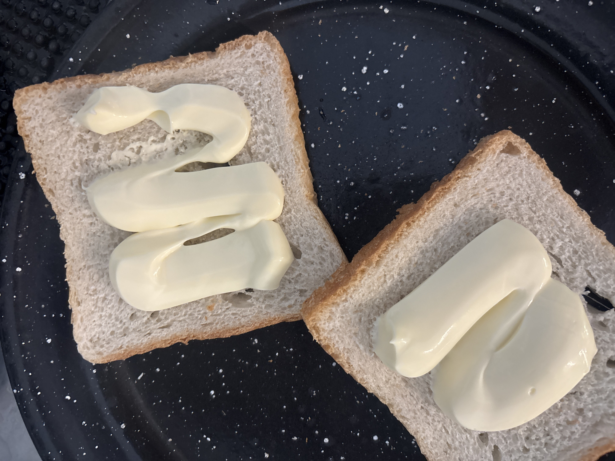 A high-angle, close-up shot reveals two slices of bread laid out on a black, speckled plate. The bread appears to be lightly toasted and rectangular. Each slice is topped with dollops of a creamy, white substance, likely mayonnaise or a similar spread. The left slice has three distinct dollops arranged in a slightly curved line, while the right slice has two dollops overlapping each other. The plate has some white specks on it, possibly remnants of food or cleaning. The image appears to have been taken indoors. The location in the city of Buenos Aires, Argentina, is unknown.