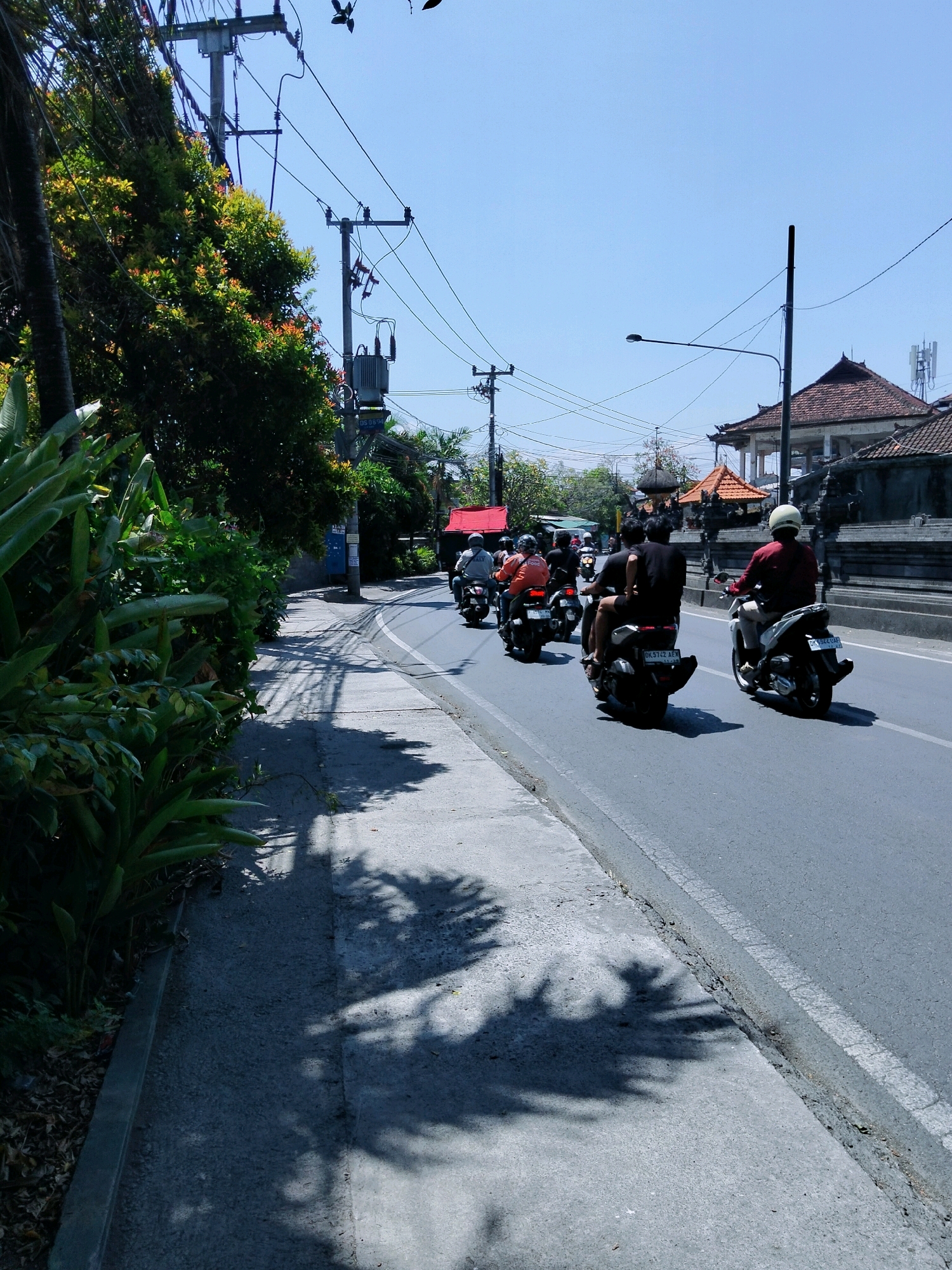 In Denpasar, Indonesia, a sunny day unfolds along a paved road lined with lush green foliage and a concrete sidewalk. The road features a white line marking its edge, and the sidewalk is shaded by the dappled shadows of trees. A procession of motorcycles, carrying several individuals, travels down the road. One motorcyclist in the foreground wears a red shirt and a white helmet, while another behind them is dressed in dark clothing. Further ahead, a motorcyclist in an orange shirt is visible, and beyond that, more riders can be seen. The background reveals traditional Balinese architecture with red-tiled roofs and ornate stone walls, suggesting the scene is in a culturally significant area. Electrical poles and wires crisscross the bright blue sky, indicating the presence of urban infrastructure. The overall atmosphere is one of daily life in a tropical city, with transportation and local architecture defining the landscape.