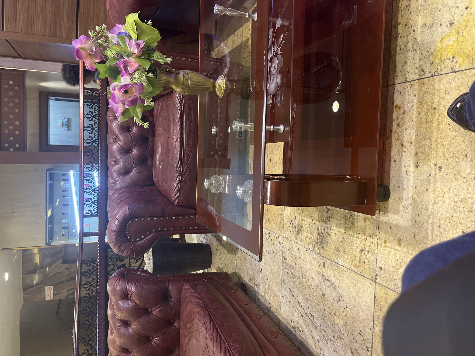 This image captures an opulent indoor waiting area or lounge, possibly within a hotel, restaurant, or business establishment in Abuja, Nigeria. The scene is dominated by two plush, deep red leather Chesterfield-style sofas or armchairs, featuring classic tufted upholstery and decorative metal studding along the arms and back.
Between the sofas sits a polished dark wooden coffee table with a clear glass top. The glass is elegantly etched with a floral design, appearing to be roses. On the table, a golden, ornate urn-shaped vase holds a vibrant bouquet of artificial flowers in shades of purple and pink, complemented by green foliage. Several decorative clear glass items, possibly small bowls or cups, are visible on a lower shelf of the coffee table.
To the left, a large mirror reflects a doorway or corridor. Within this reflection, a white sign with "Ladies" and "Gents" text is visible, indicating restrooms. The reflection also shows what appears to be a person with dark, curly hair. Further back, another glass partition or display cabinet reflects bottles or items, suggesting a bar or retail area. A "No Smoking" sign is partially visible through a glass panel on the left.
The floor is covered with light-colored, speckled granite or marble tiles, featuring various shades of beige, grey, and yellow. In the lower right corner, a person's right leg and foot, wearing dark trousers/jeans and a dark sandal, are visible, suggesting someone is seated or standing nearby. The overall ambiance is one of sophisticated comfort.