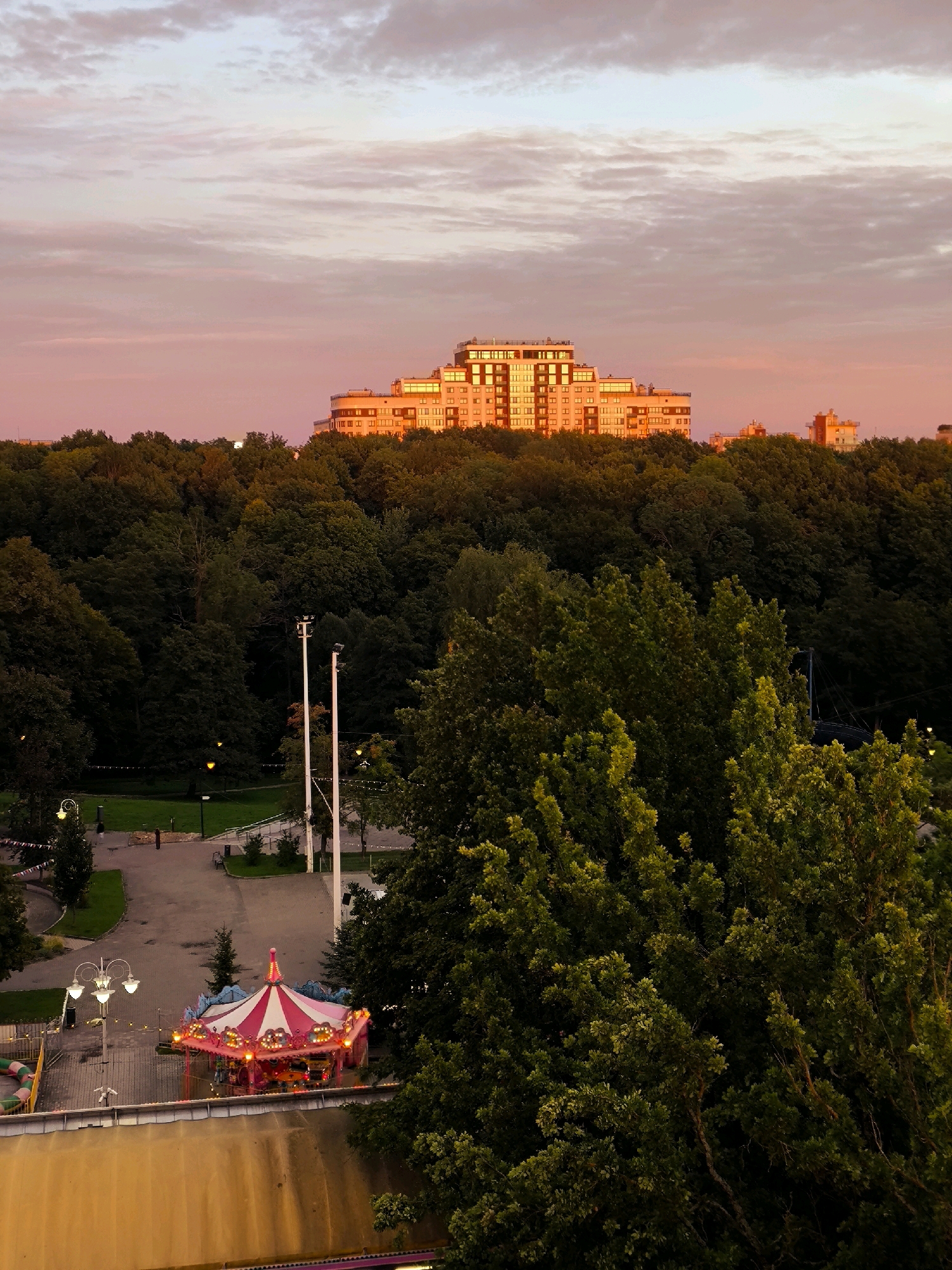 The image depicts an aerial view of a park in Kaliningrad, Russia, during sunset. The sky is filled with pink and purple clouds, illuminated by the setting sun. In the background, a large, multi-story building with orange and white facades stands out, its windows reflecting the warm glow of the sunset. The foreground is dominated by a dense forest of green trees, with a small carousel visible in the lower left quadrant. The carousel is brightly lit with pink and white lights, and its striped canopy suggests it is part of an amusement park. A paved path leads through the park, with lampposts and benches along the way. The overall scene evokes a peaceful and serene atmosphere.