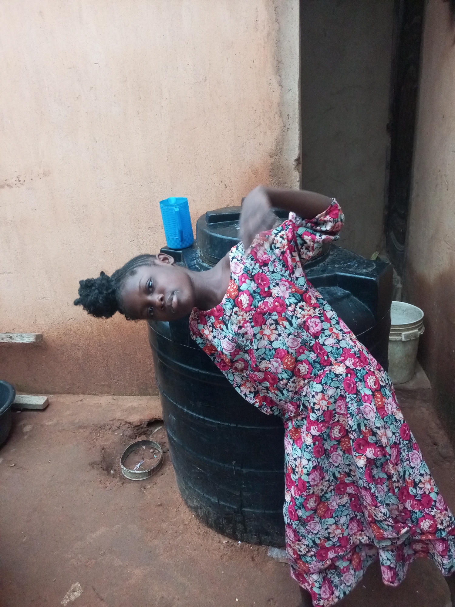 A young Black female is posing by a large black plastic water tank. She is wearing a long-sleeved, floral-print dress with red, pink, and teal flowers. Her hair is styled in a bun on top of her head. The tank is positioned on a dirt ground outside a building with a tan, textured wall. To the left of the tank, a blue plastic container is visible. Further to the right, a white bucket sits near the building. The environment suggests a rural or semi-urban setting within Tempure, Nigeria, likely a residential compound given the presence of domestic water storage and basic building materials. The lighting indicates it is daytime.