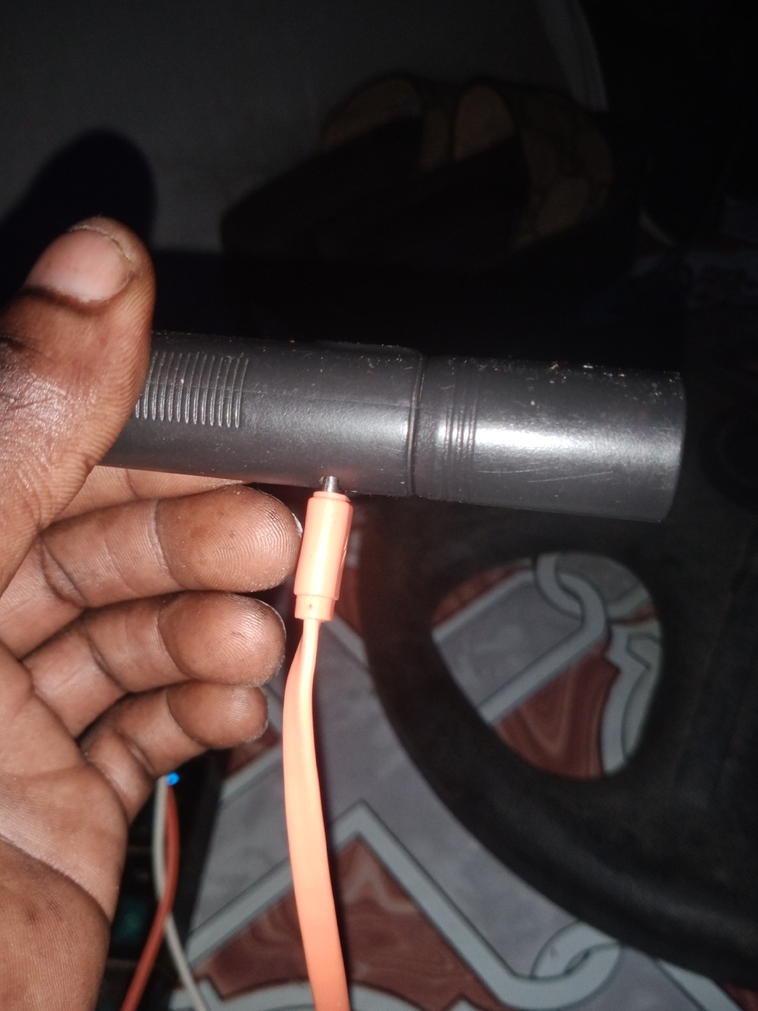 A close-up shot shows a hand holding a gray cylindrical object, possibly a microphone or a flashlight, with a ribbed texture near one end. An orange cable with a metallic connector is plugged into a small port on the object. The hand is dark-skinned, with visible fingerprints on the fingers. The background is dark and blurry, with a hint of a patterned surface beneath the object. The lighting suggests the image was taken indoors in low light conditions. The presence of what appears to be a charger or cable indicates the object might be electronic. The location of this scene is unclear but could be a residence or a workshop in Jalingo, Nigeria.