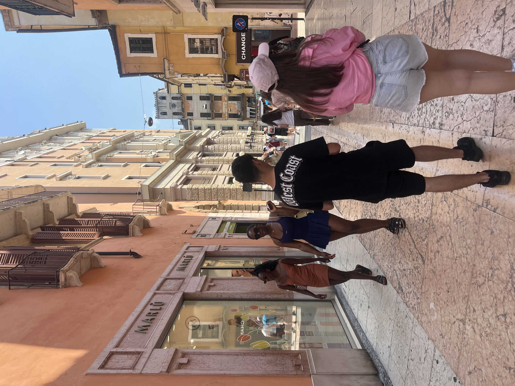 A street scene with pedestrians and buildings in Verona, Italy. The image is oriented vertically.
In the foreground, a group of people walk on a stone-paved street. Two women, one in an orange dress and the other in a navy dress, are walking past a store with the name "MARLO" displayed prominently above its storefront. A man in a black shirt with "West Coast" printed on the back is walking in the same direction, followed by a woman in a pink jacket and denim shorts.
Behind the people, the street extends into the distance, lined with tall buildings. The architecture of the buildings is characteristic of old European cities, with ornate detailing and a variety of window styles. A "CHANGE" sign is visible in the background, indicating a currency exchange service. The sky is a clear blue, suggesting a sunny day. Other pedestrians can be seen walking in the background.