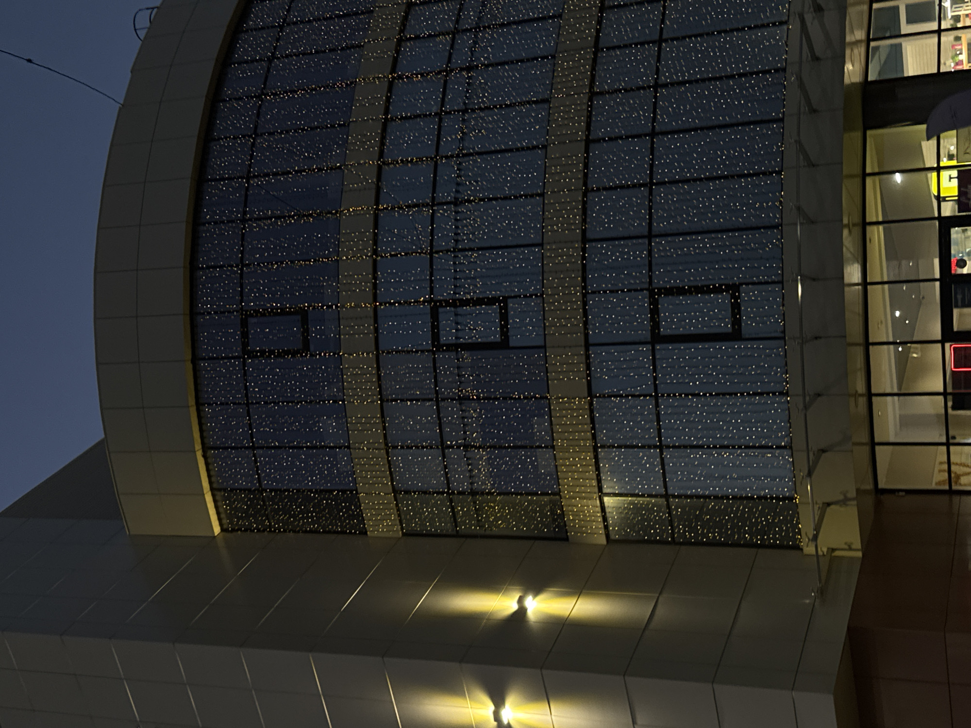 A modern building with a curved glass facade decorated with strings of fairy lights stands against a dark blue sky. Two upward-facing lights illuminate the grey tiled exterior wall on the left side of the building. The glass facade is segmented by horizontal bands of yellow and is divided into multiple panes by dark vertical mullions, creating a grid-like pattern. Each pane is filled with a dense arrangement of small, twinkling white lights that create a starry effect. The overall impression is of a building decorated for a festive occasion or an evening event. The lower portion of the building shows a ground-level entrance with large glass doors and illuminated interior spaces.
