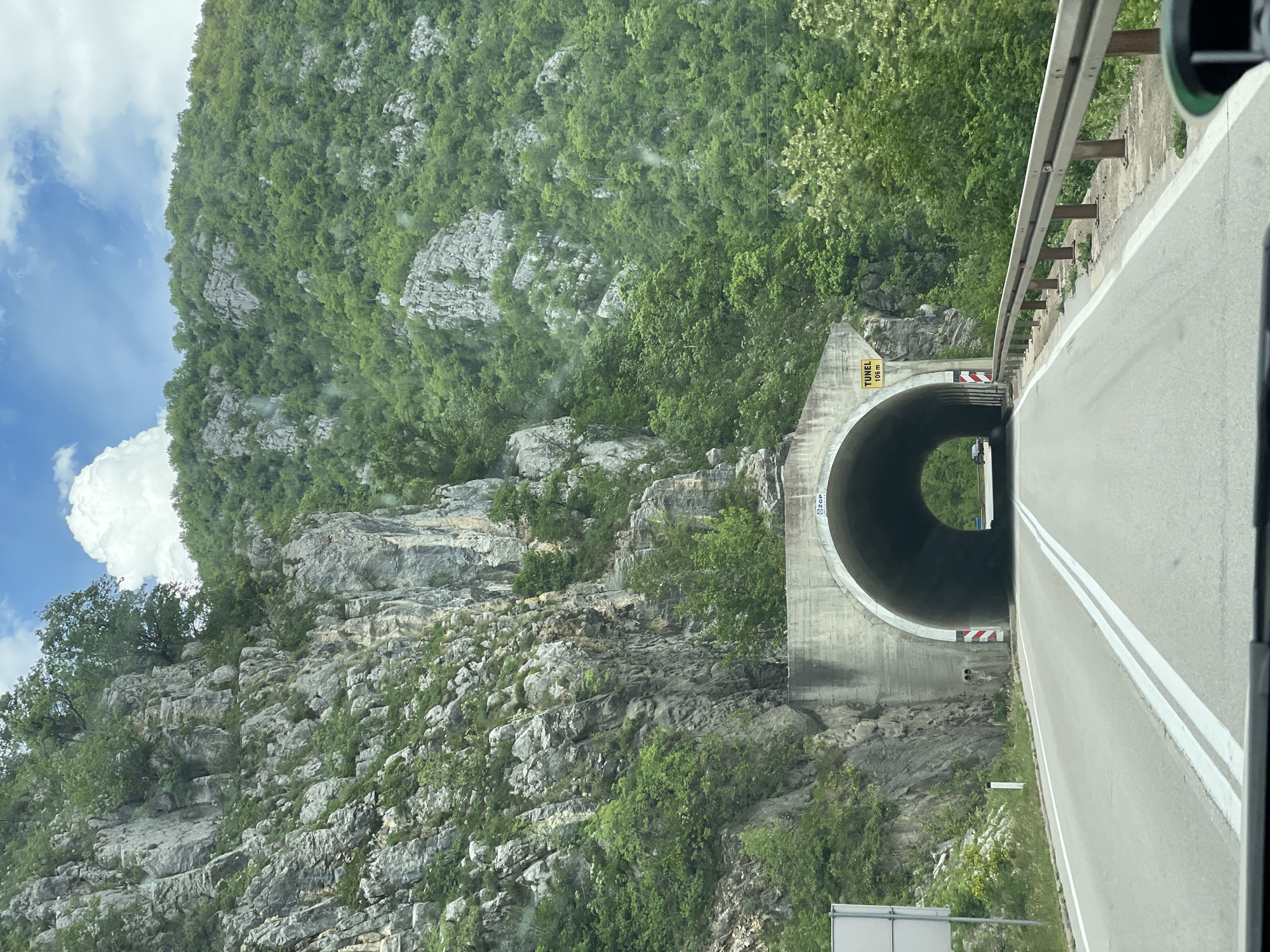 The image captures a road leading into a concrete tunnel in Zdaljevac, Bosnia and Herzegovina. The road is paved with double white lines marking the lanes. A metal guardrail is visible on the right side of the road. The tunnel entrance is surrounded by a rocky, tree-covered mountain. A yellow sign with "TUNEL 108 m" is visible above the tunnel entrance. The sky is partially visible on the left, showing clouds.