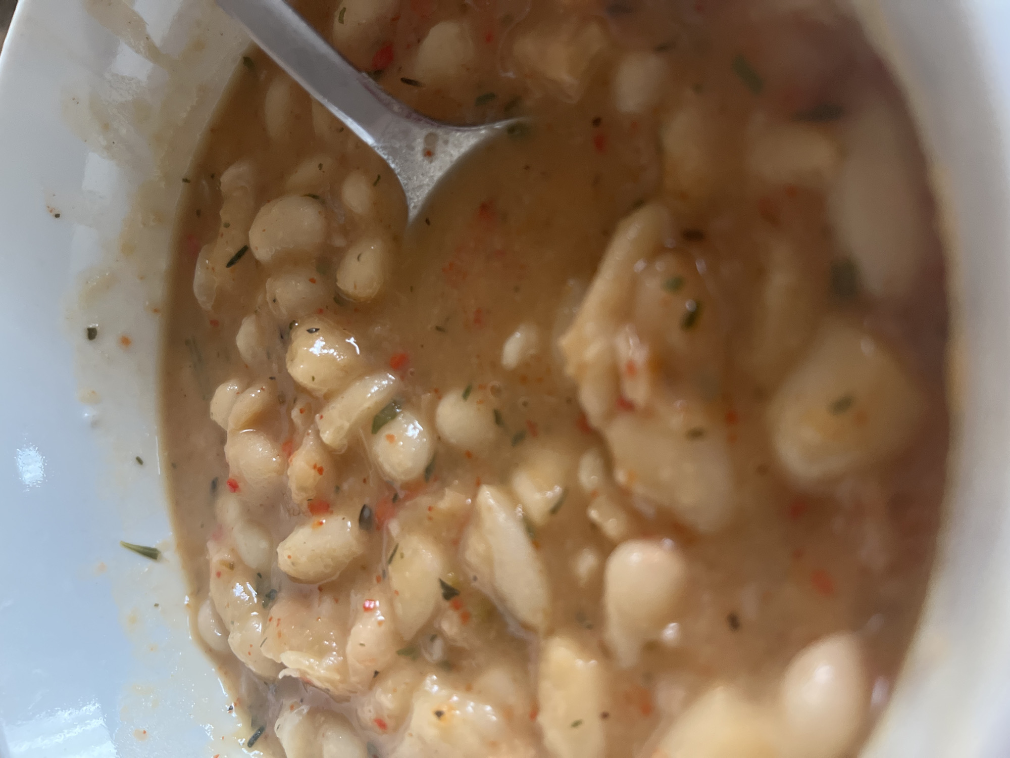 A close-up, overhead shot shows a white bowl filled with a bean and broth-based soup or stew. A silver spoon is partially submerged in the soup. The beans are white and of varying sizes. The broth is a light brownish color with flecks of green and red spices. The bowl has a light interior.