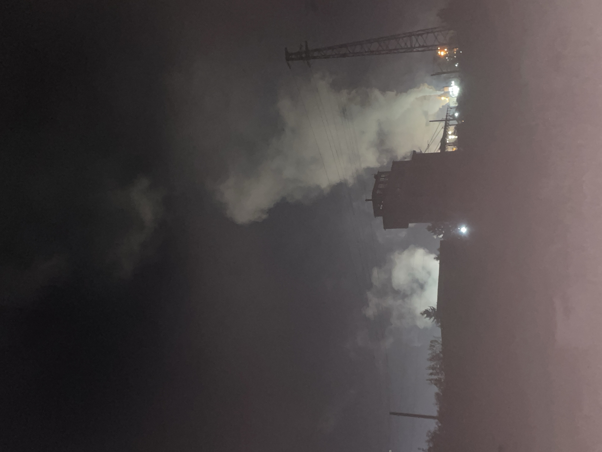 The image depicts a nighttime scene in Razgrad, Bulgaria. A tower or tall building stands in the middle, and a metal transmission tower is visible in the upper portion of the frame. The sky is filled with clouds or fog, illuminated by distant city lights. Electrical lines run horizontally. A street light is visible below a building. Trees line the lower right portion of the scene.