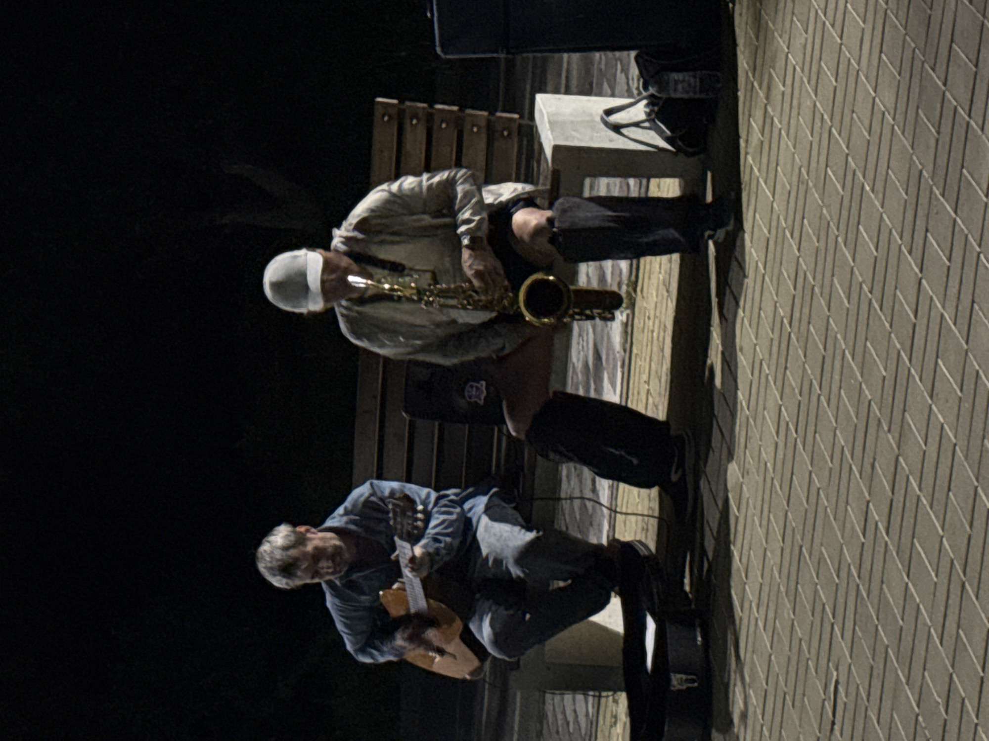 Two musicians are performing on a park bench at night. The person on the left is playing an acoustic guitar and wearing a blue shirt and jeans. The person on the right is playing a saxophone and wearing a white cap and a light-colored jacket. Both are sitting on a wooden bench in what appears to be a public park. The ground in front of them is paved with interlocking bricks. To the right of the saxophone player, a black speaker is visible, and a black bag sits on the ground near the bench. The background is dark, suggesting it is nighttime. The scene is likely in Pavlodar, Kazakhstan, given the provided context.