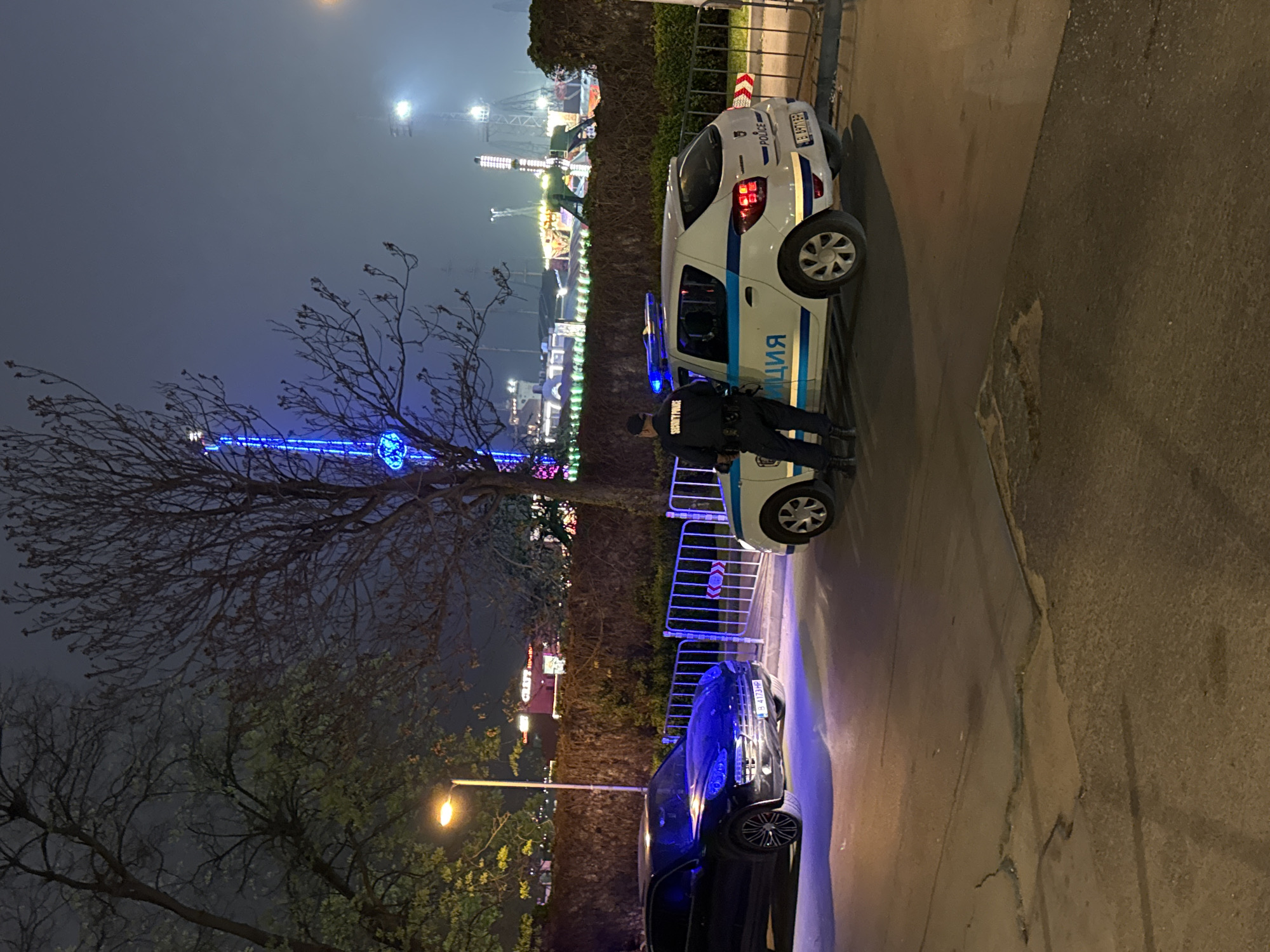 The image shows a nighttime scene in Varna, Bulgaria. In the background, a fair or amusement park is illuminated with colorful lights. In the foreground, a white police car is parked, with an officer standing beside it. The word "ПОЛИЦИЯ" (Police) is written in Cyrillic on the side of the car. A black car is parked behind the police car. A bare tree branch extends into the frame from the left side. Metal barriers are placed in front of the vehicles. The scene is illuminated by streetlights and the lights from the fair. The ground is paved.