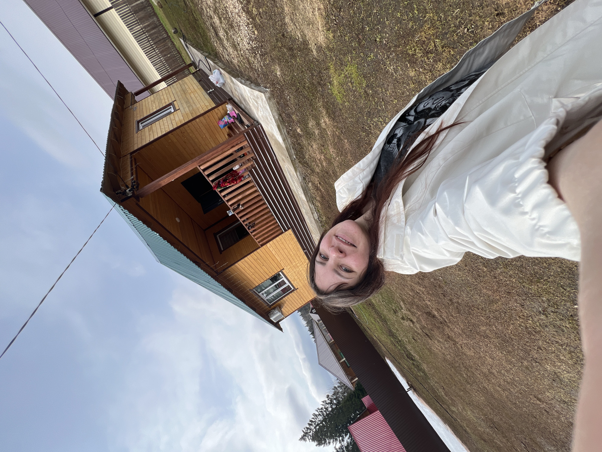 A woman with long brown hair, wearing a white jacket over a black and white patterned top, is taking a selfie outdoors. Behind her is a two-story wooden house with a green metal roof. The house has a wooden balcony and a staircase leading up to it. To the left of the house, the sky is visible with wispy clouds. To the right is an area of dry grass. The location is identified as Bol'shakino, Russia. Other buildings with metal roofs are partially visible in the background. A power line stretches across the upper left of the frame.