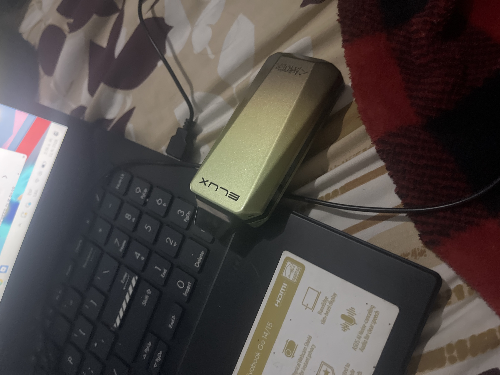The image depicts a close-up, angled view of a laptop keyboard with a gold-colored electronic device plugged into it. The laptop appears to be a dark color, and some of its keys are visible, including "P", "Shift", "End", and numerical keys. The electronic device is rectangular with rounded edges and has the text "ELUX" and another unreadable marking on its surface. A black cable extends from the device and disappears off-frame. The background consists of a textured surface with a pattern of brown and beige shapes, likely a blanket or bedding. The lighting is dim, suggesting an indoor environment. No specific location in Santa Ana, Venezuela can be determined from the provided image.