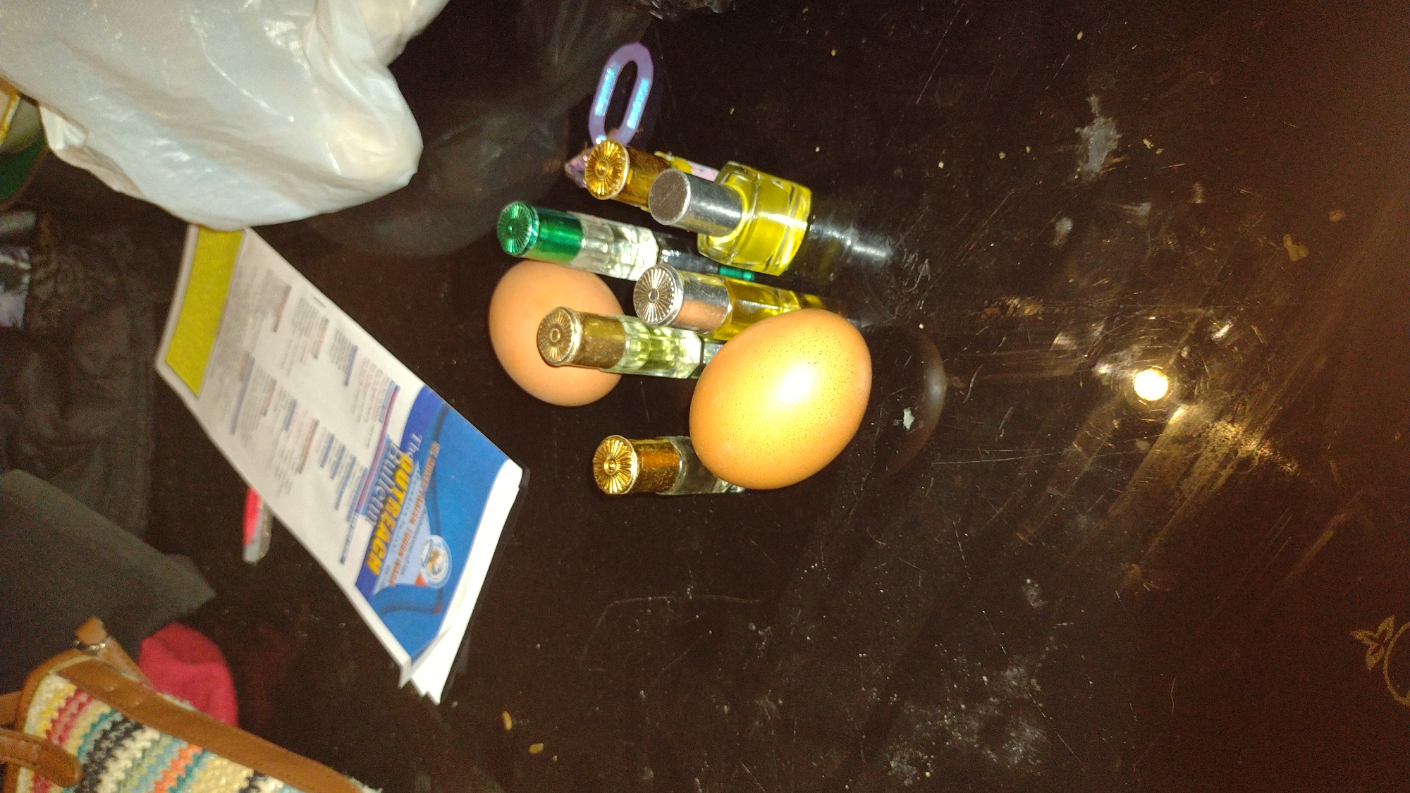 A still life composition on a dark, reflective surface features two brown eggs and six small glass perfume bottles. The bottles vary in shape and cap color, with some featuring gold caps and others silver or green. One bottle is square, while the others are cylindrical. Positioned behind the eggs and bottles is a folded piece of paper with text and graphics, appearing to be a bulletin or flyer. To the left, a portion of a colorful woven bag with brown leather straps is visible. A white plastic bag is partially visible on the right side of the frame. The scene is lit by an overhead light source, casting reflections on the glossy surface and highlighting dust and scratches. The location is a table in Jos, Nigeria.