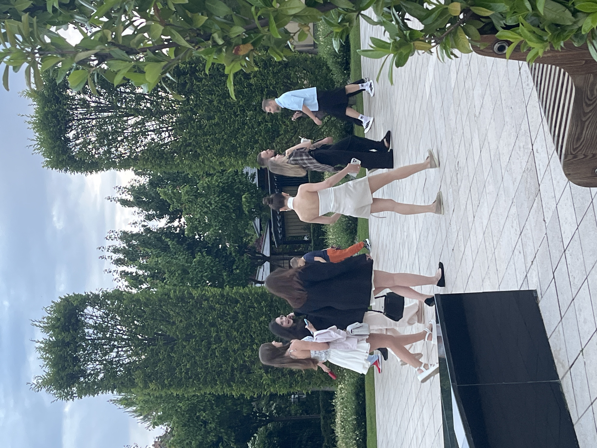 This image captures an outdoor social gathering on a light-colored tiled patio, surrounded by abundant green trees and foliage. The sky above is overcast or partly cloudy, indicating daytime, possibly late afternoon given the diffused light.
Several individuals, mostly young adults, are visible, standing and mingling. In the foreground, a group of women includes one in a black coat over a light dress, another in a white dress with a patterned top section, and a third woman, partially seen in a light dress, holding a phone. Behind them, a child in orange pants and a dark top can be observed.
Further into the scene, a woman with dark hair tied back wears a white backless top and holds a phone. Close by, another woman in a plaid shirt and dark pants holds a beverage. A man with a beard is also present, dressed in a light blue polo shirt, dark shorts, and sneakers. He appears to be holding a phone.
The ground is a patterned tiled surface. To the right, a dark, rectangular block-like structure is in the foreground, and a wooden bench with vertical slats is partially visible further back, near the top. The dense greenery suggests a garden or park-like setting. The scene is set in Krasnodar, Russia, based on the provided context. There is no visible text in the image.