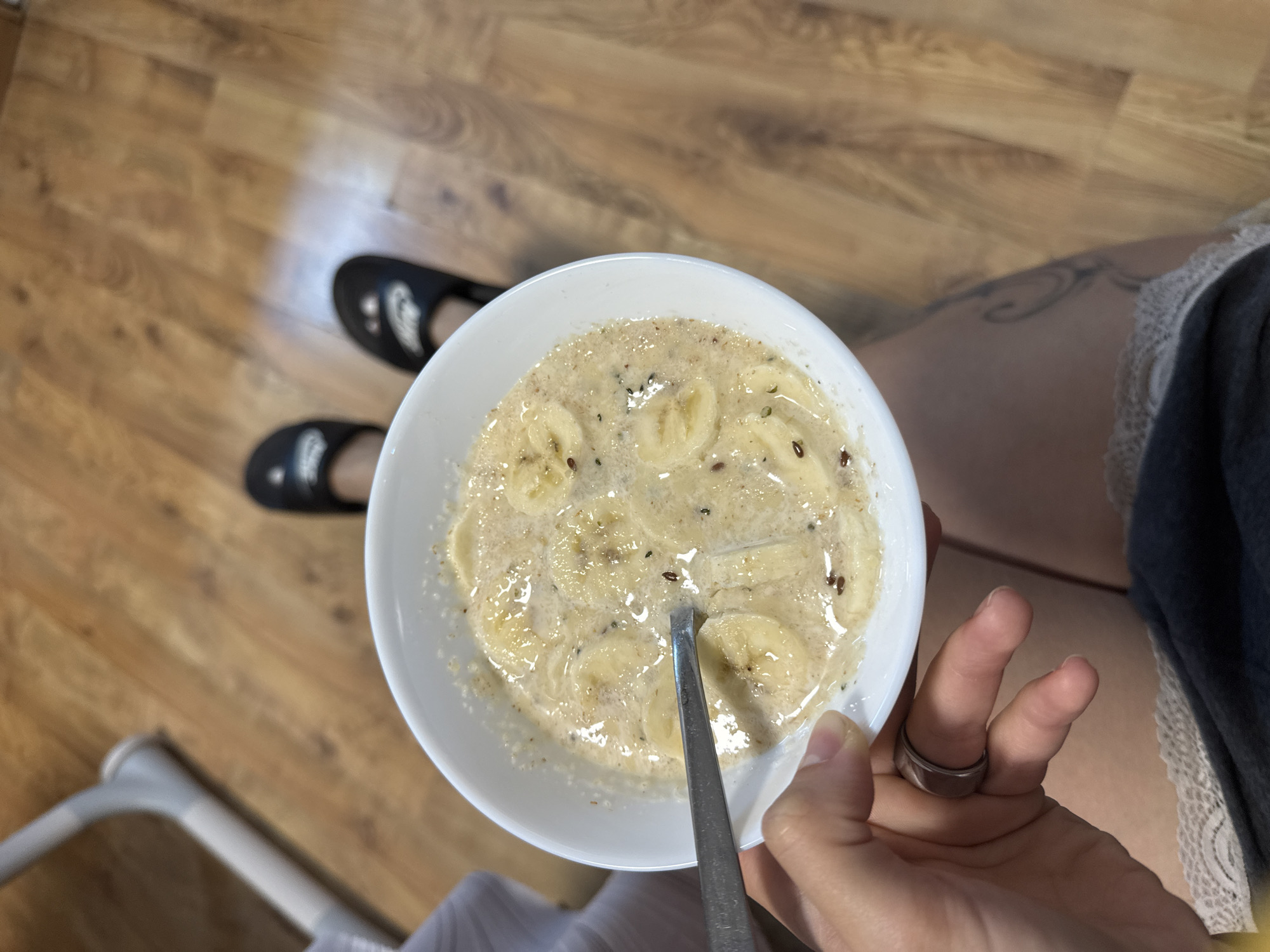 In an indoor setting with a wooden floor, a person's hand holds a white bowl filled with oatmeal topped with sliced bananas and flax seeds. A silver spoon is partially submerged in the oatmeal. The person is wearing dark slides on their feet. The background shows a portion of a high chair and the wooden floor.