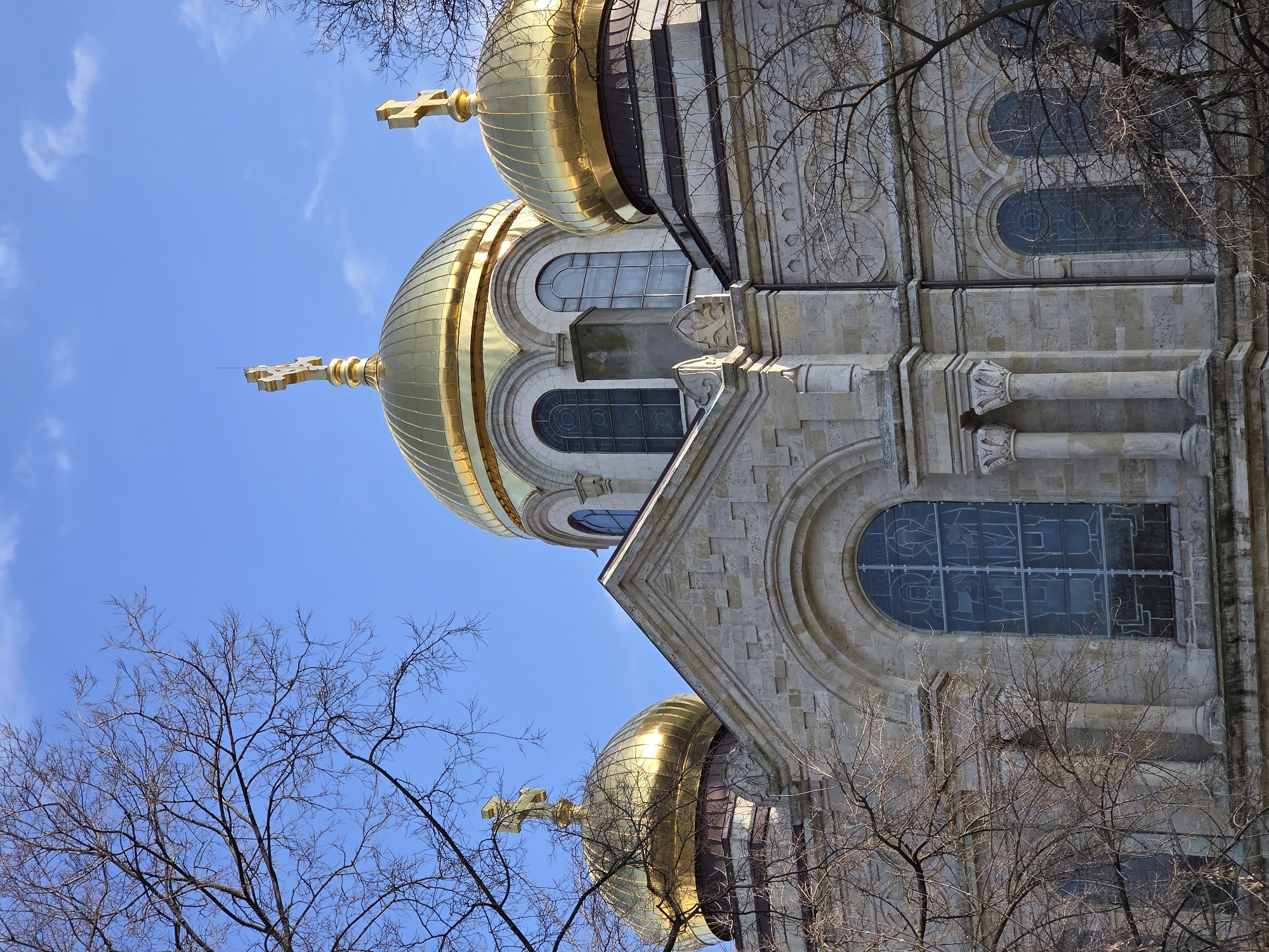 The image displays the upper sections of the Dormition of the Theotokos Cathedral, located in the city center of Varna, Bulgaria, facing Saints Cyril and Methodius Square. The primary subjects are the cathedral's light-colored, carved stone facade and its prominent golden onion domes.
Three major golden domes are visible, each topped with a gold Orthodox cross. The largest central dome is positioned towards the upper middle of the frame, flanked by a smaller dome on the right and another partially visible dome on the far left. A fourth gold Orthodox cross is visible on the far left, suggesting another dome beyond the frame. The domes are ribbed, reflecting bright sunlight. A lightning rod is visible atop the central dome.
The stone facade features arched windows, decorative pilasters, cornices, and carved details. Several arched windows are discernible; a large, tall arched window on the lower left displays a stained glass pattern depicting two schematic figures. Other arched windows, including a pair on the central dome's structure, are also visible, featuring patterned glass panes. A narrow, dark-colored chimney or vent stack is situated between two windows on the central section.
Bare, deciduous tree branches, devoid of leaves, partially obscure the cathedral's facade on the left and lower right sections of the image. The sky above is clear and bright blue, with scattered, wispy white clouds. No people, specific actions, or interactions are present in the scene. The lighting indicates daylight, with direct sunlight illuminating the domes and parts of the facade.