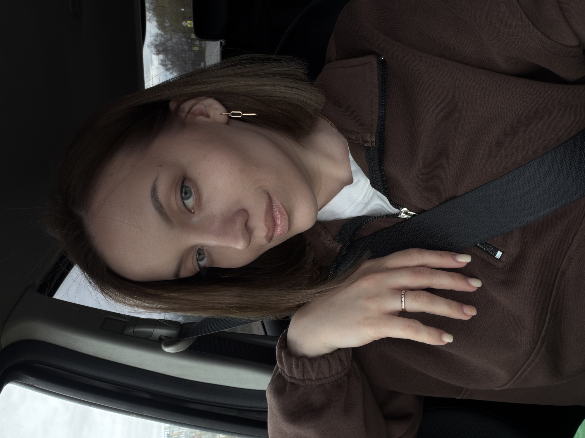 A woman is photographed inside a car, likely in Слобода, Russia. She wears a brown zipped sweatshirt, a white t-shirt underneath, and a gold ring on her left hand. A gold linear earring is visible on her left ear. She is wearing a seatbelt. The background visible through the car window suggests an urban environment.