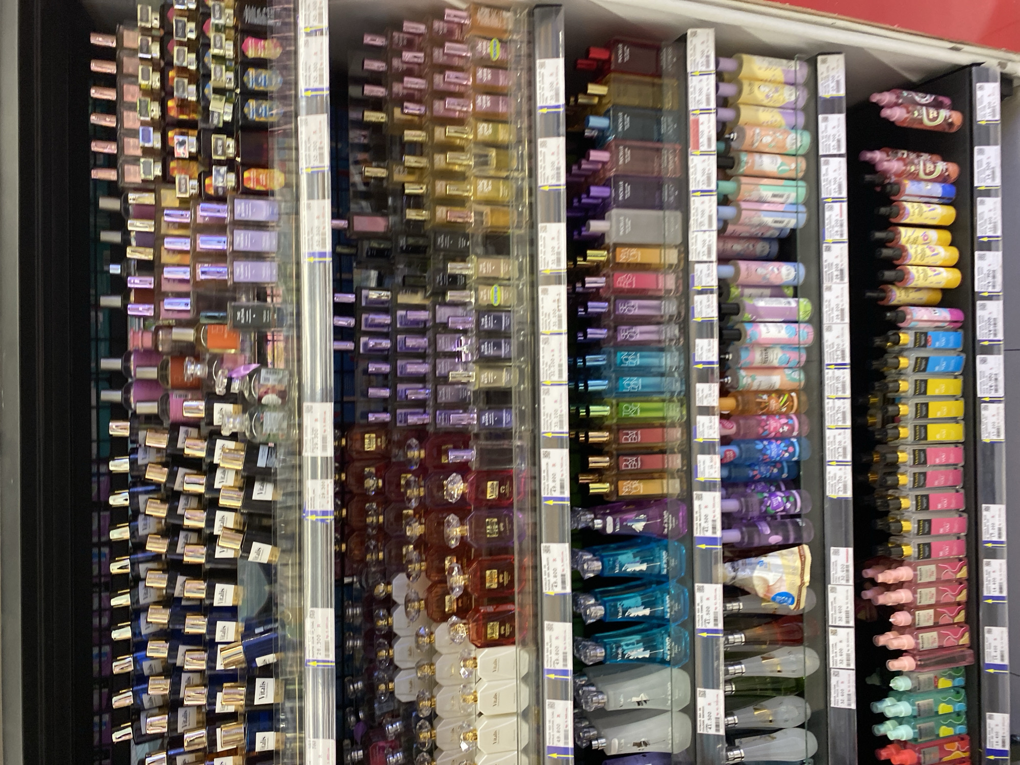 The image shows a retail shelf display filled with various perfumes and body mists. The products are arranged in rows on multiple shelves. The perfumes are mostly in clear glass bottles of various shapes and sizes, containing liquids of different colors including blue, pink, purple, yellow, and clear. Many of the bottles have gold or silver caps. Some of the bottles have labels with text, though it is largely illegible. The products are neatly organized. The overall environment appears to be a well-lit retail store. The image was taken in Banda Aceh, Indonesia, at a store, likely a department store or a specialized beauty store, given the extensive display of fragrance products. There are no people visible in the image.