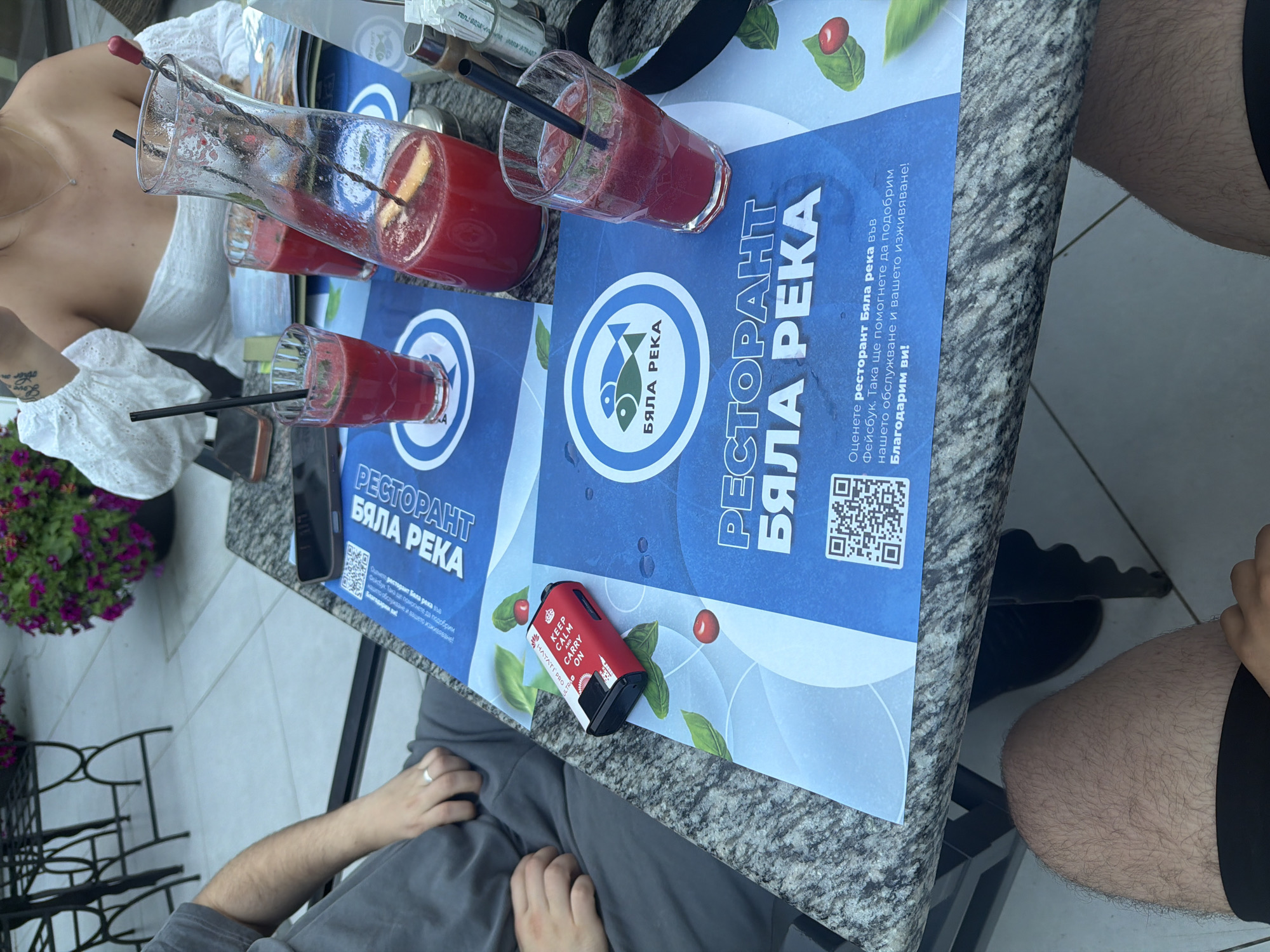 The image shows a table at an outdoor restaurant, likely in Kalofer, Bulgaria, given the text on the placemats. In the foreground, a person's legs are visible, wearing black shorts. On the table, there are two glasses filled with red juice, a pitcher of the same red liquid, and a black electronic cigarette device with the text "KEEP CALM AND CARRY ON". The placemats on the table feature the logo and name of a restaurant called "БЯЛА РЕКА" (Byala Reka), which translates to "White River". The background shows part of a woman wearing a white top, and decorative plants in a pot. The setting appears to be a pleasant outdoor dining area, possibly a patio or terrace.