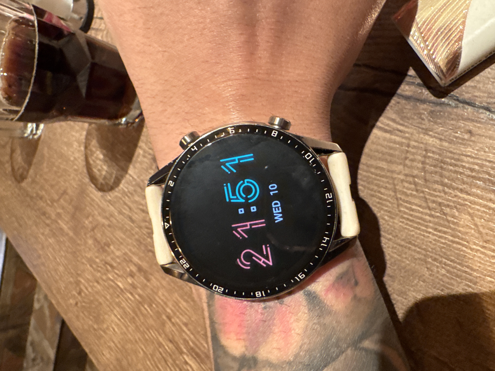The image shows a close-up of a person's wrist wearing a smartwatch. The smartwatch has a black face with digital time displayed as "21:51" in pink and blue digits. Below the time, it reads "WED 10". The watch has a white strap and a metallic bezel with markings for hours. The background is a wooden surface, likely a table, and a glass of dark liquid, possibly soda or beer, is visible in the upper right corner. The lighting suggests it is daytime. There is no visible indication of the specific location in Varna, Bulgaria.
