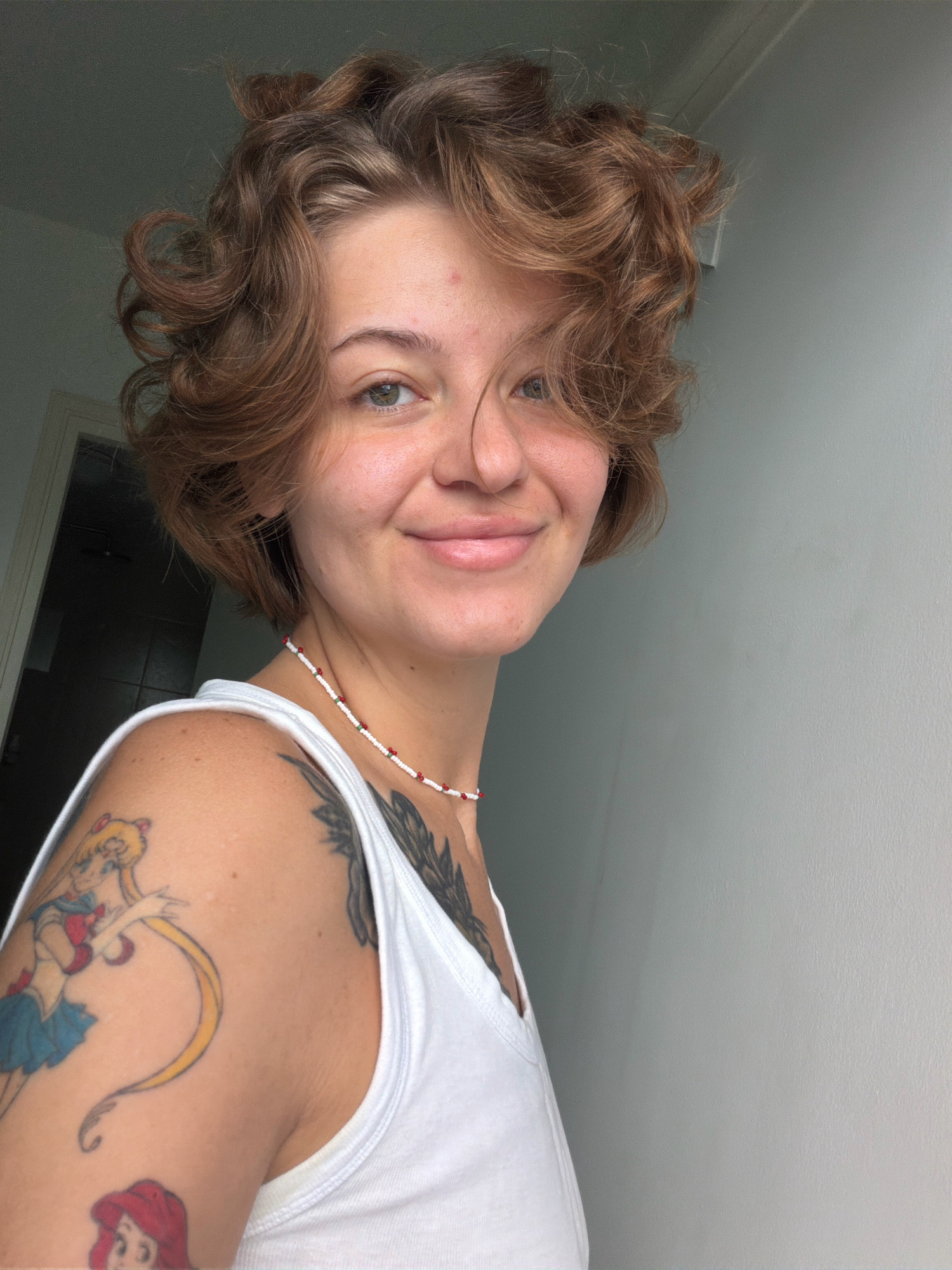 A young woman with short, curly brown hair and light green eyes smiles directly at the camera. She is wearing a white tank top and a beaded necklace with red and white beads. She has tattoos on her left arm and shoulder, including one of Sailor Moon. The background is a plain, light gray wall. The image appears to have been taken indoors, possibly in a residential setting, and there is no indication of the specific location within Hua Hin City Municipality, Thailand.