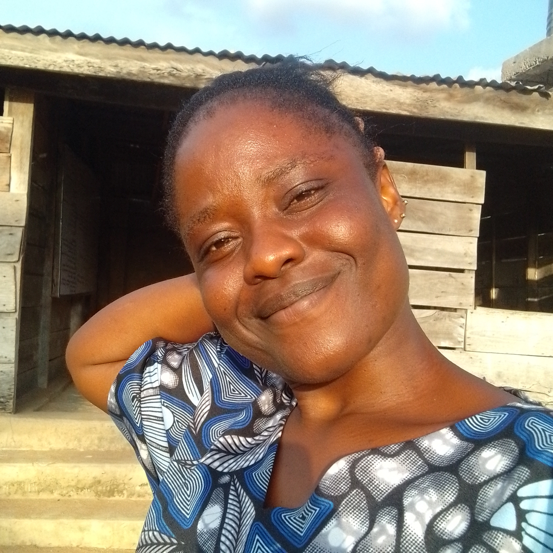 This is a close-up selfie of a dark-skinned woman with a warm smile, taken outdoors in Jalingo, Nigeria. She is resting her left arm behind her head, and her right arm is out of frame. She is wearing a blue and white patterned top. Her hair is dark and pulled back. The background shows a weathered wooden structure with a corrugated metal roof, suggesting a simple, possibly rural, setting. The time of day appears to be late afternoon or early evening, as indicated by the warm sunlight casting shadows and highlighting her skin. The sky is partly cloudy. There is no visible text in the image. The woman's relaxed posture and joyful expression convey a sense of contentment and ease.
