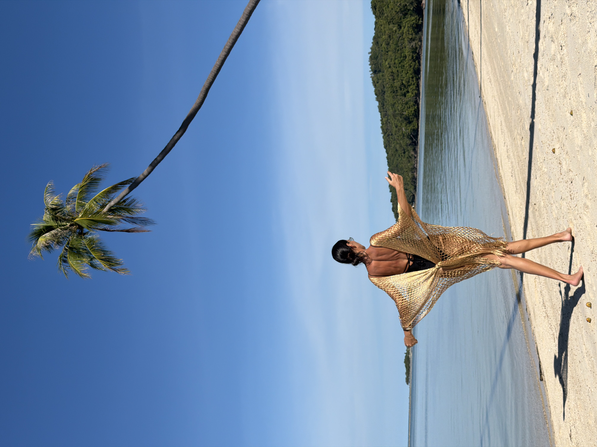 The image depicts a person standing on a sandy beach with their back to the camera, facing the ocean. The person is wearing a gold, lacy cover-up over a black swimsuit. They are looking towards the right side of the frame, with their left arm extended upwards and outwards. To the left of the person, a palm tree trunk curves dramatically against a clear blue sky, with its fronds visible at the top left corner. The ocean stretches out in front of the person, with a lush green forest visible in the distance along the coastline. The sun appears to be high in the sky, casting a distinct shadow of the person and the palm tree onto the sand. The overall impression is one of a serene and sunny day at a tropical beach. The image is oriented in landscape mode, with the horizon running vertically down the center. The location is identified as Baan Thong Krut, Thailand. There is no visible text in the image.