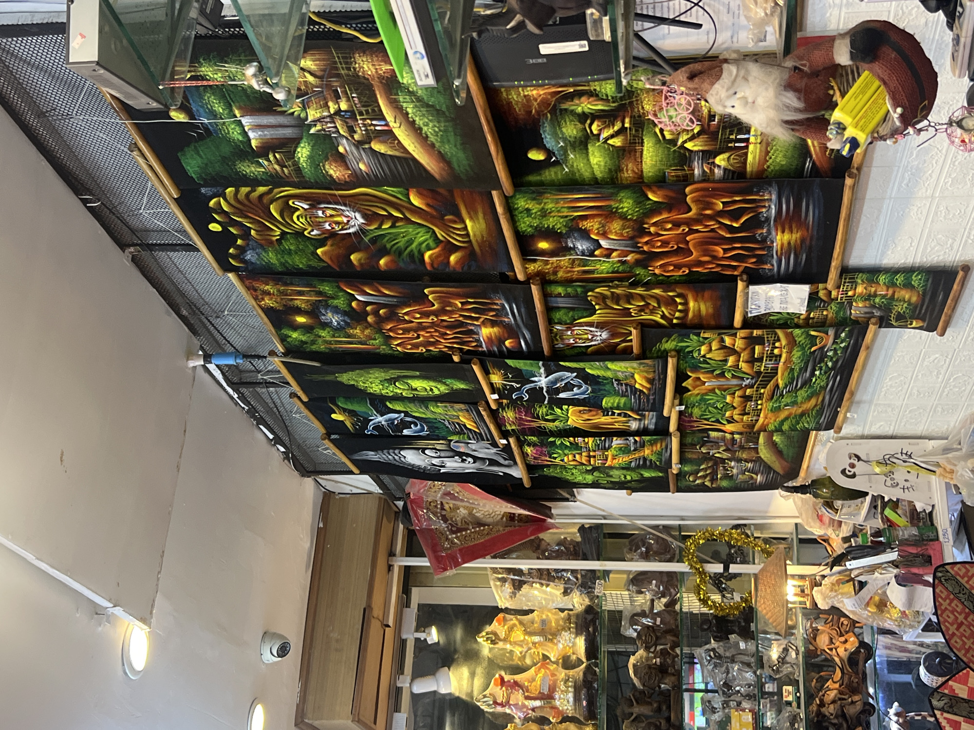 This is a medium-shot, eye-level interior photograph of a market stall in Karon, Thailand. The stall displays numerous large, colorful paintings on fabric, depicting various scenes of nature and animals such as tigers and horses, as well as Buddhist iconography. The paintings are hung vertically, supported by wooden dowels. In the foreground, on a shelf, there are decorative items, including a lamp with a woven shade, bottles, and various small trinkets. A plush Santa Claus figure is hanging from a hook on the right side of the frame. Behind the paintings, a security camera is visible on the ceiling, as are light fixtures. The overall impression is of a busy souvenir shop or market stall offering a variety of artistic goods.