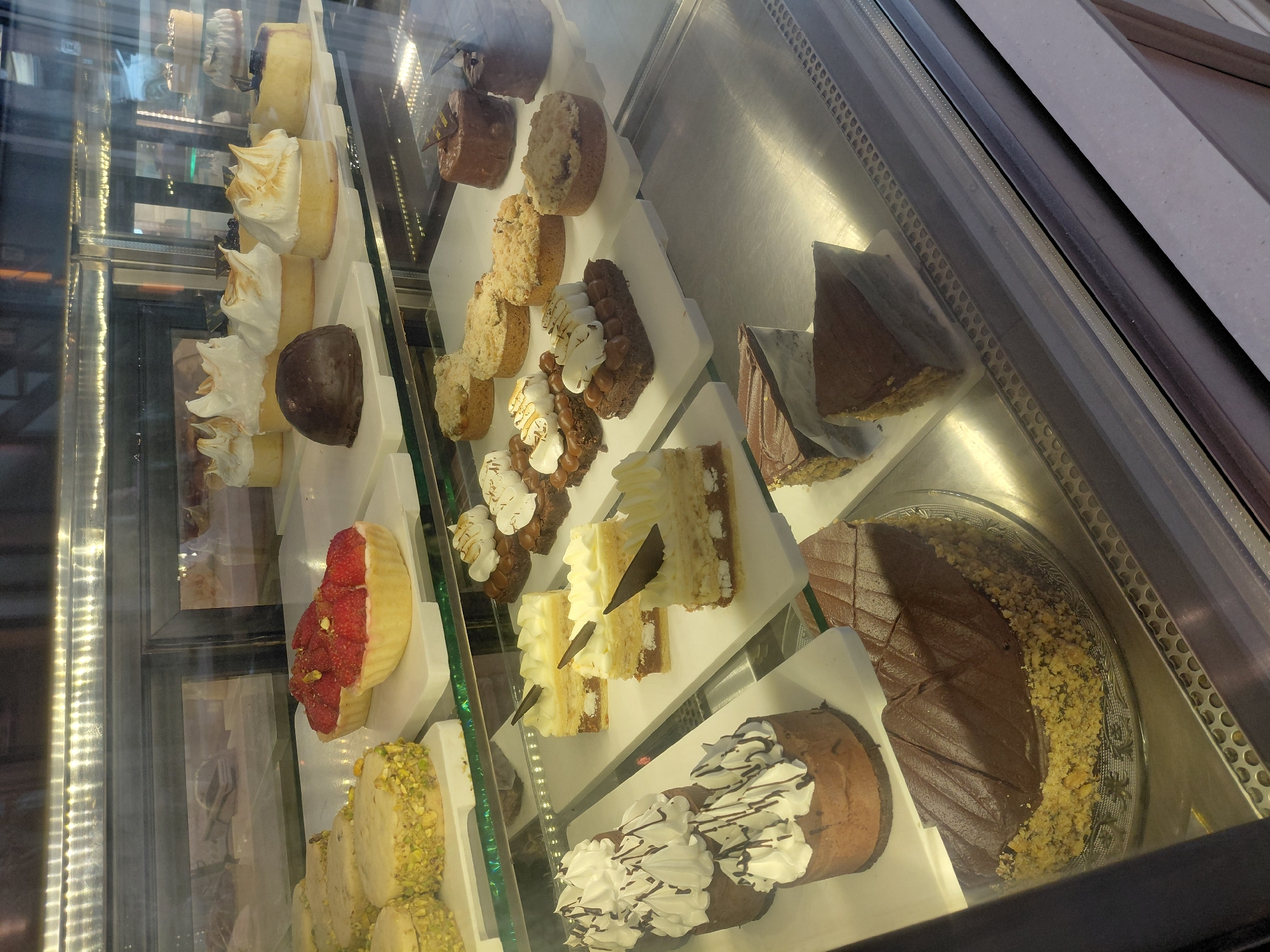 Inside a refrigerated display case, likely in a bakery or cafe in Ushuaia, Argentina, are various desserts. From left to right: slices of cake with meringue topping, a round chocolate mousse, various other cakes with fruit toppings or nuts, and a large chocolate cake decorated with nuts around the edge. The display case has glass on multiple sides, creating reflections. The environment is brightly lit. There are no people visible in the image.