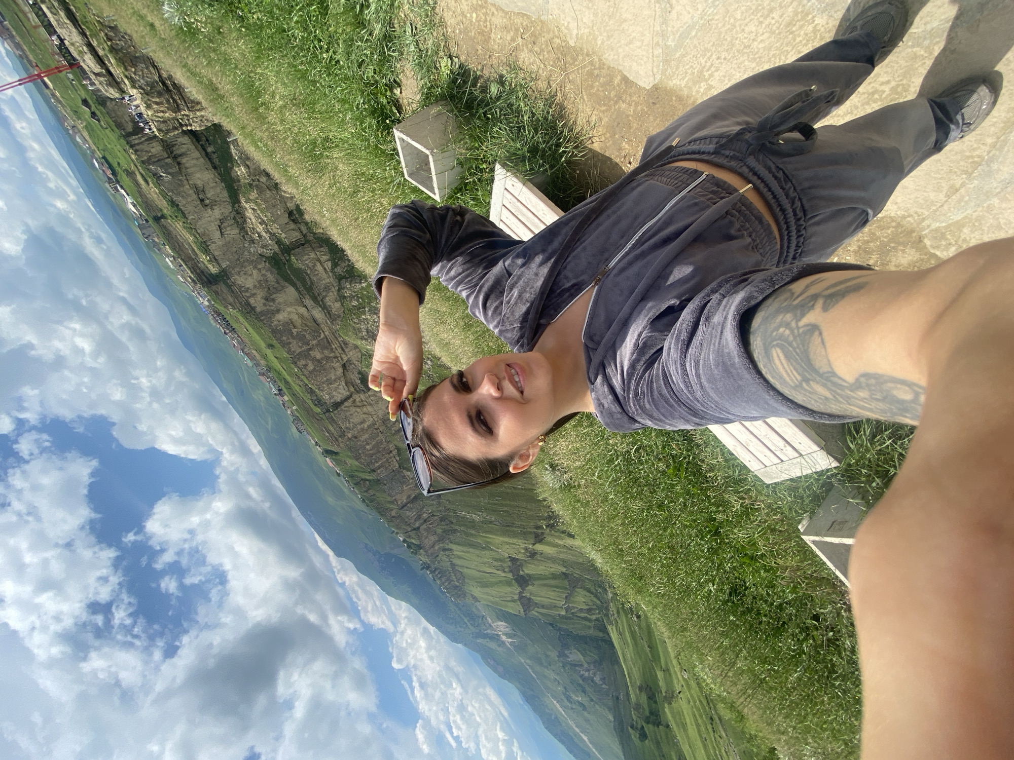 A woman is taking a selfie outdoors, with a vast mountainous landscape in the background. She is wearing a grey velour tracksuit and sunglasses on her head. She is standing on a grassy slope next to a wooden bench and a concrete planter. The background features a dramatic canyon with a village nestled along its edge, under a cloudy sky. The location appears to be the village of Арани, Russia, known for its high-altitude scenic views.