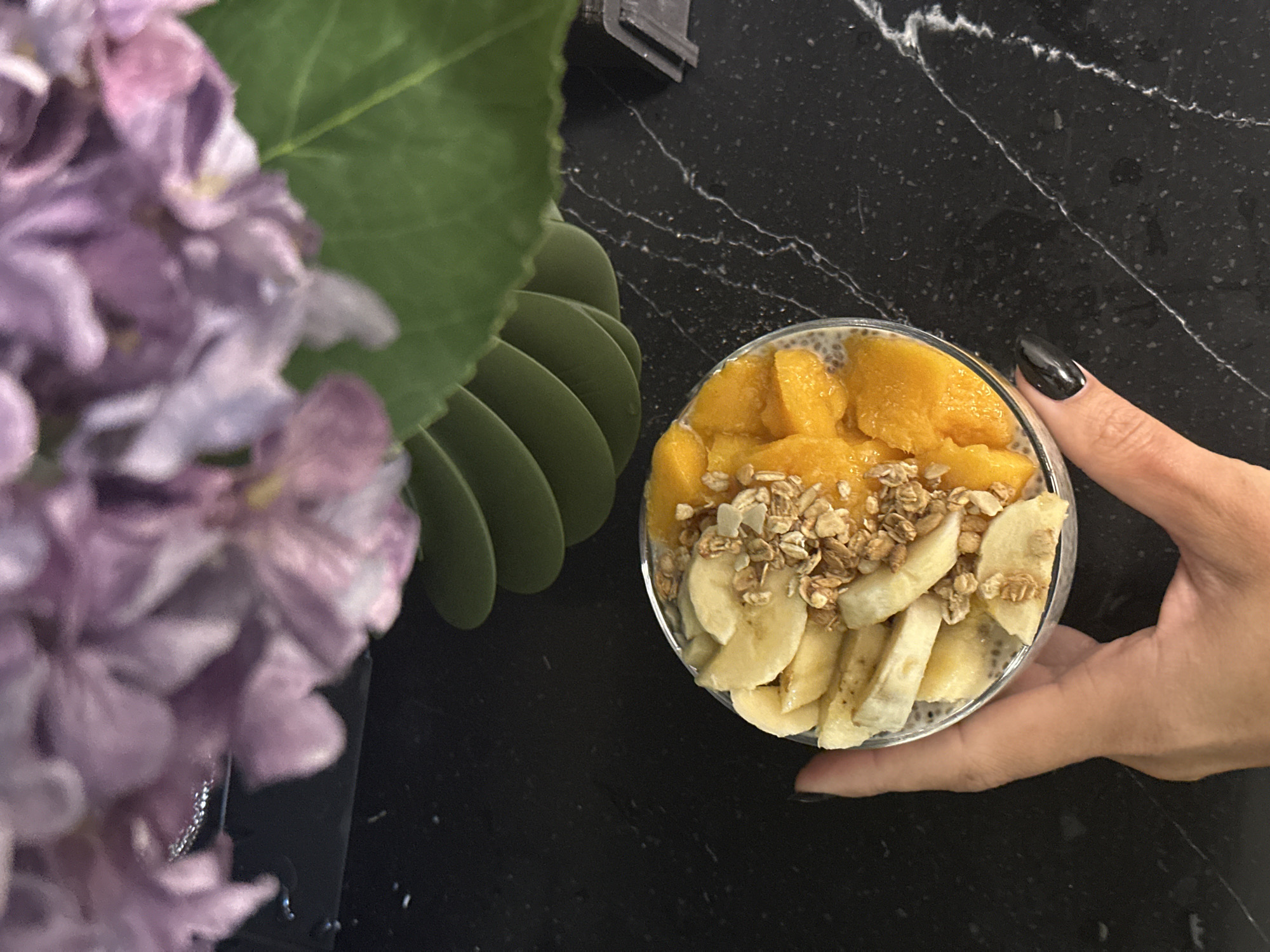 A well-lit, overhead shot displays a dessert bowl, likely a chia seed pudding, topped with banana slices, diced mango, and granola, held by a hand with dark nail polish. To the left, out-of-focus purple flowers with green leaves add a decorative element. The scene is set on a black marble countertop. No indication in the image suggests that the location is Novosibirsk, Russia. The overall impression is of a healthy snack presented in a domestic environment.