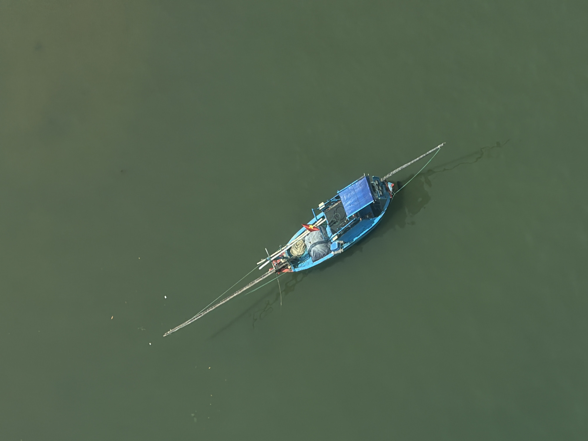 The image is an aerial shot of a single blue fishing boat floating on the surface of green water. The boat has a sunshade structure, possibly made of canvas, at the stern. A red and yellow flag is visible on the boat. Long fishing poles or nets extend from both the front and back of the boat. There are some lines or ropes trailing in the water. The water is calm and relatively uniform in color. The location is Nha Trang Ward, Vietnam.