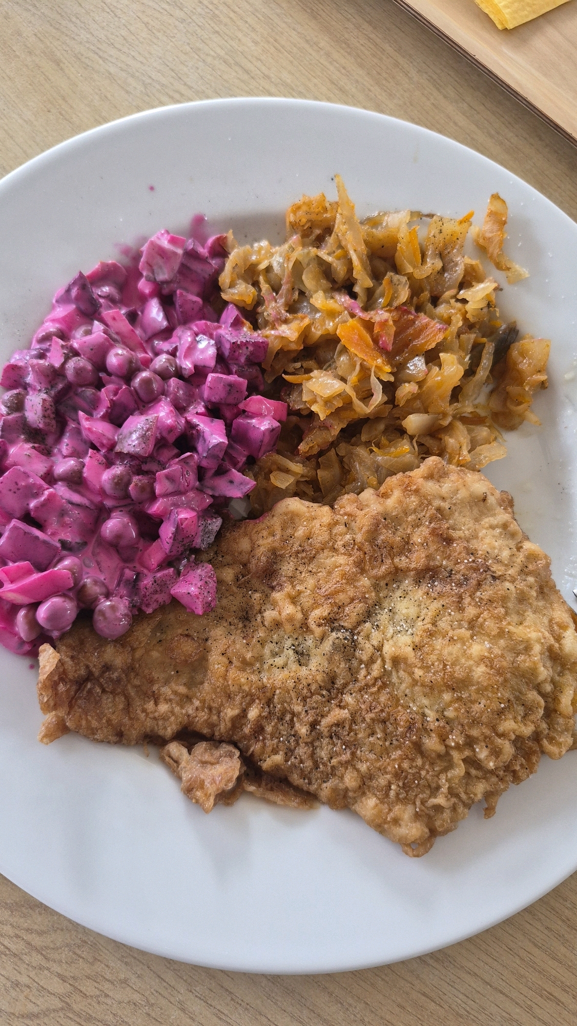 A close-up, overhead shot shows a white plate filled with food, placed on a wooden table. The plate contains a large, breaded and fried piece of meat, a pile of cooked cabbage, and a vibrant pink beet salad. The beet salad is mixed with what appears to be peas and a creamy dressing, with visible diced beets and peas. The cooked cabbage is a mixture of shredded cabbage and possibly carrots, with a slightly translucent appearance. The meat cutlet is golden brown and seasoned with visible black pepper. The wooden table has a light brown, grainy texture. A small portion of a yellow object, possibly cheese, is visible on a separate surface in the upper right corner of the image, suggesting it might be a serving platter or a continuation of the table setting. There are no people visible in the image, and no specific location in Ludza, Latvia can be identified from the visual information provided.