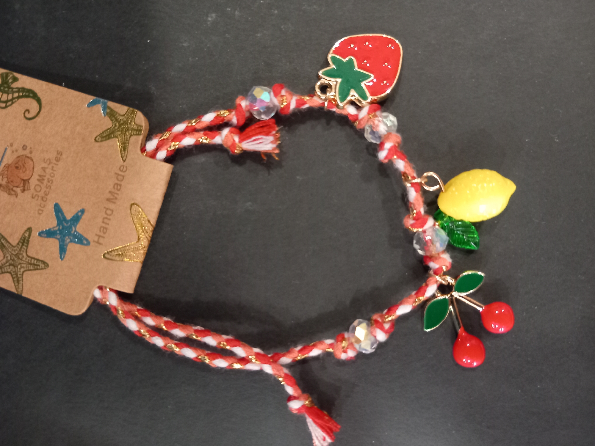 A close-up, eye-level shot shows a handmade bracelet with fruit charms against a dark, textured surface, possibly a table. The bracelet is made of red and white braided string with gold thread woven in. It is adorned with several charms: a red strawberry with green leaves, a pair of red cherries with green leaves, and a yellow lemon. Clear, multifaceted beads are also incorporated into the bracelet's design. A light brown cardboard tag with nautical-themed graphics (starfish, seahorse, whale) and text like "SOMAS accessories" and "Hand Made" is visible at the top of the image, suggesting the bracelet is new or for sale. The lighting is somewhat harsh, creating shadows and highlights. The overall impression is of a craft item with a summer or beach theme.