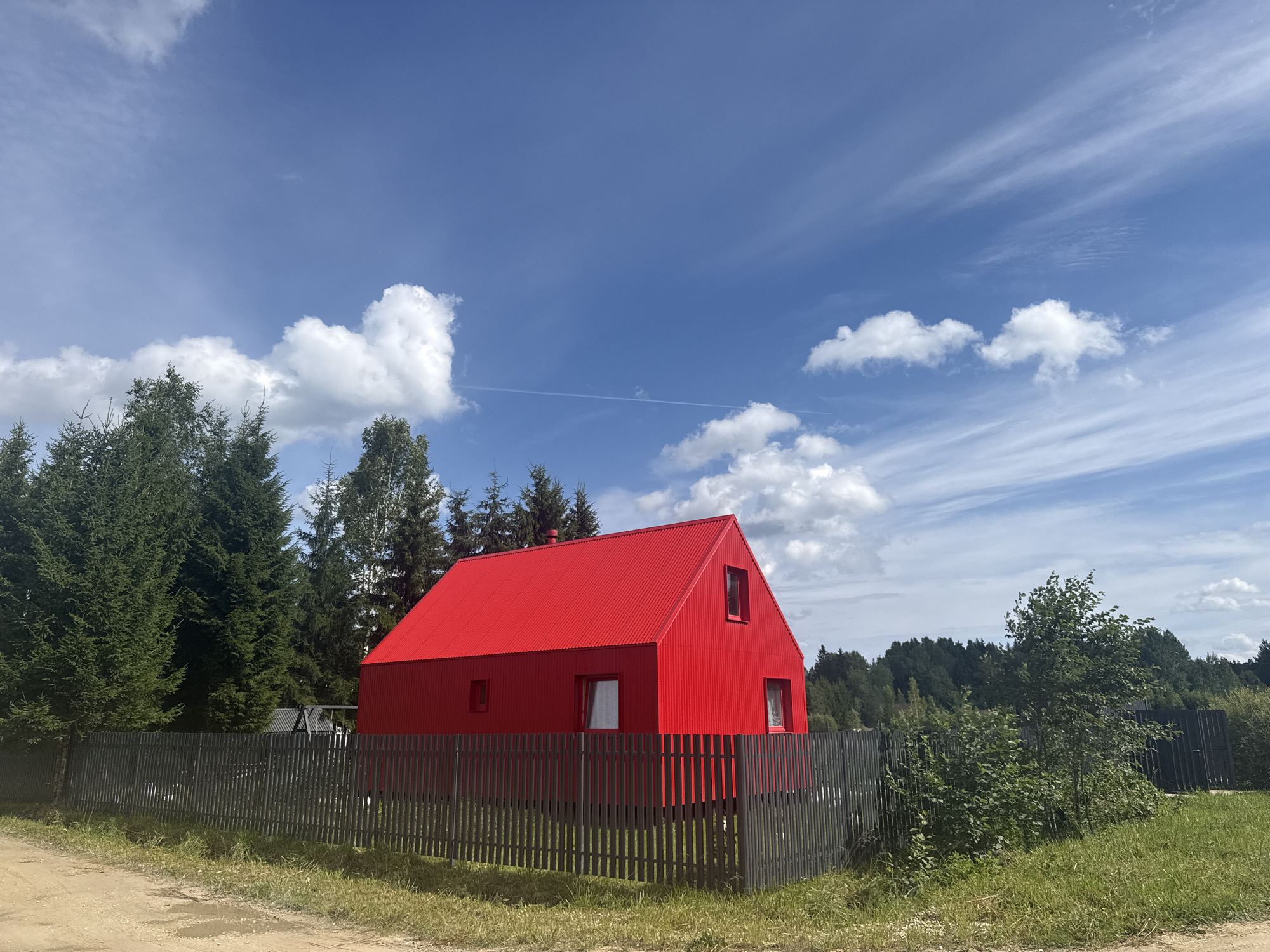 The image depicts a bright red, modern, angular house situated in a rural setting. The house has a corrugated metal exterior and a steep, red, corrugated metal roof. There are at least three windows visible on the front and side of the house, one of which is a triangular dormer window on the roof. The house is surrounded by a dark gray, vertical slat fence. To the left of the house, a dense line of dark green coniferous trees is visible. The background consists of a lush green landscape with rolling hills covered in trees, indicating a natural or forested environment. The sky is a vibrant blue with scattered white, wispy clouds, and a faint contrail from an aircraft is visible. The foreground is a dirt road with some patches of grass and puddles, suggesting a recent rain or damp conditions. The image appears to have been taken on a sunny day with good visibility. The location is identified as the city of Lyady, Russia. There are no people or animals visible in the image.