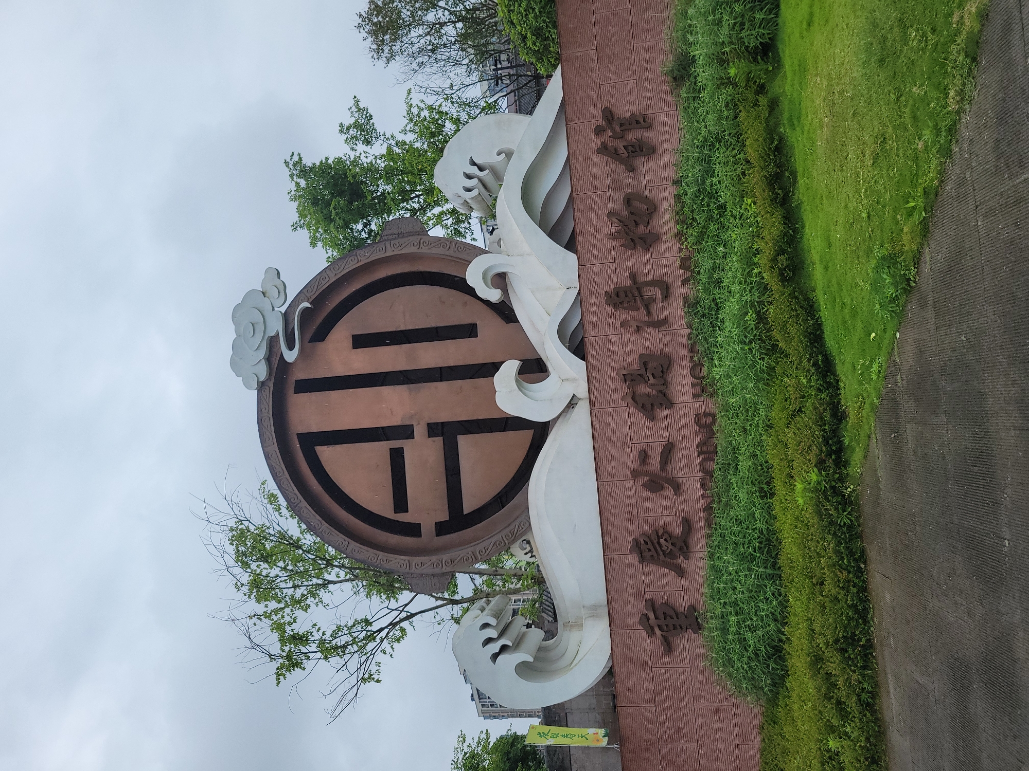 The image depicts a large sign for Southwest University in Hechuan, China. The sign features a circular copper-colored emblem with an ornate design. White stylized waves flank the emblem. The Chinese characters are vertically arranged on a reddish-brown textured background. Green plants and grass grow along the base of the sign. The sky is overcast. A sidewalk or paved area is visible on the right side. Trees are present in the background.