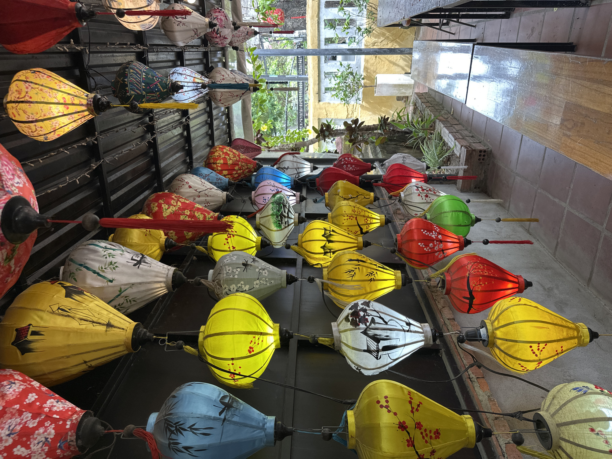 This is a close-up, eye-level shot of a vibrant display of colorful fabric lanterns hanging from a ceiling. The lanterns are varied in size and design, featuring patterns of flowers, bamboo, and traditional Asian scenery. They are illuminated from within, casting a warm glow. The lanterns are predominantly in shades of red, yellow, blue, white, and green. Some lanterns have decorative tassels. The ceiling appears to be made of corrugated metal. In the background, a brick wall is visible, with some greenery and parts of outdoor seating, including wooden benches and tables with metal legs. The scene is set outdoors, likely in a courtyard or covered walkway. The atmosphere suggests a cultural or tourist-oriented establishment in Hội An Ward, Vietnam, known for its lantern tradition. There are no people visible in the image.