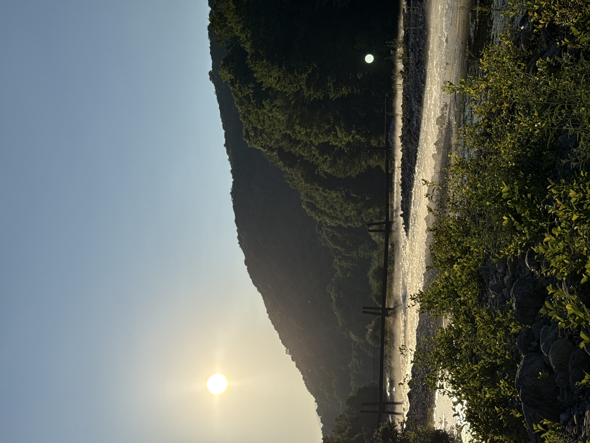 The image shows a natural landscape with a prominent sun low in the sky, indicating either sunrise or sunset. The sun is a bright, glowing orb casting warm light across the scene. In the foreground, dense green foliage grows alongside a rocky embankment, suggesting a natural, perhaps mountainous or hilly, environment. A body of water, possibly a river or a lake, flows or stretches through the scene, its surface reflecting the sunlight, creating a shimmering, dynamic effect.
In the middle ground, a dark, tree-covered mountain or hillside slopes steeply towards the water. The vegetation on the hillside is thick and appears lush, catching the sunlight in places and creating a textured, shadowed appearance. A dark structure, possibly a bridge or some form of infrastructure, spans across the water, with visible supports. The presence of this structure, along with what appears to be a light fixture on the hillside in the background, suggests human presence or development within the natural setting. Another bright light, possibly a reflection or artificial light, is visible on the dark hillside in the distance.
The overall mood of the image is serene and picturesque, illuminated by the warm, golden light of the sun. The clear, pale blue sky further enhances this feeling of peace and natural beauty. The image appears to be taken in a location with significant natural features, with the vegetation and the sun's position suggesting a temperate climate. No text is visible in the image.