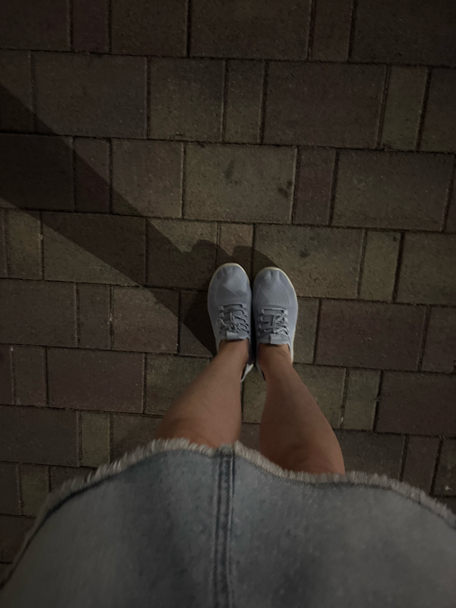 The image shows a person's legs and feet from a high angle, standing on a brick pathway. The person is wearing light blue sneakers and a denim skirt with a frayed hem. A shadow from an unseen object falls across the pathway to the left of the person's feet. The lighting suggests it is nighttime or a dimly lit indoor space. There is no visible indication that this scene is in Sochi, Russia.
