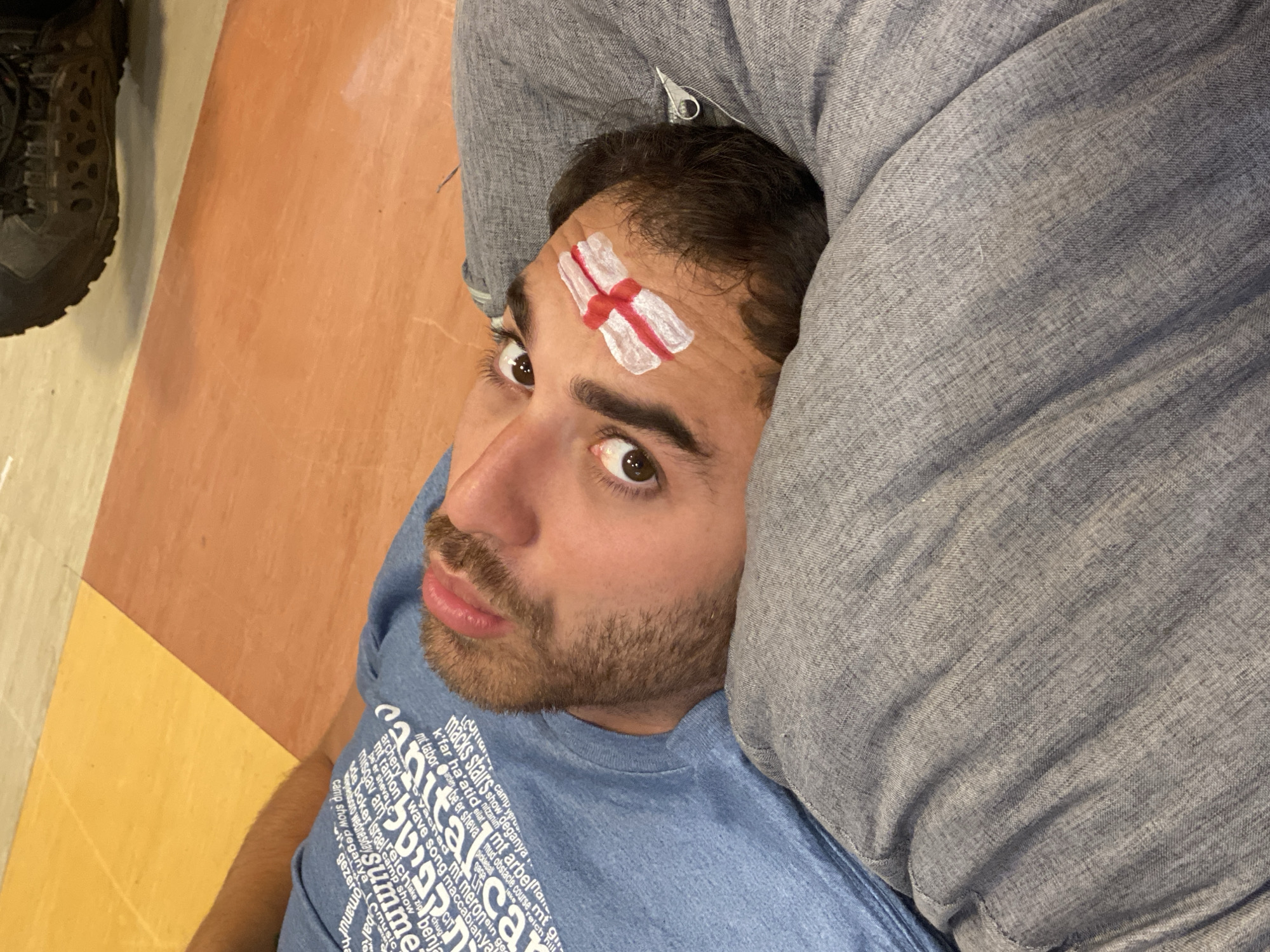 A young man lies on his back, his head tilted to the right. He has a light complexion, a beard, and brown eyes. His right eyebrow is accentuated with an England flag painted in white and red. He is wearing a blue t-shirt with white text printed on it. He appears to be lying on a person wearing gray pants. The background consists of a floor with yellow and orange tiles. The scene is likely indoors, possibly at a sporting event or gathering. There are no discernible landmarks that would place the image specifically in Washington Township, United States of America.
