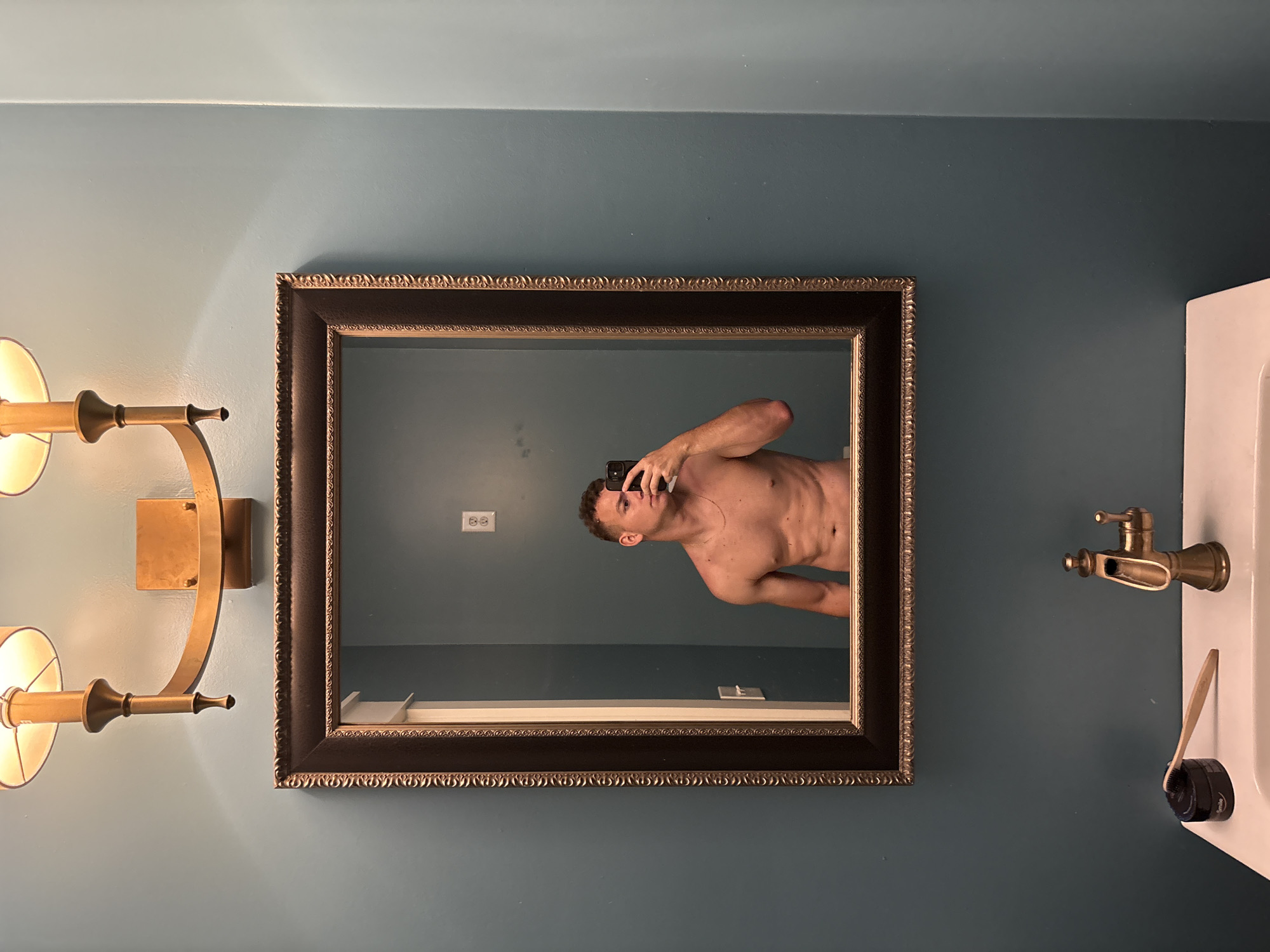 This image depicts a bathroom scene. A shirtless man is taking a mirror selfie in a well-lit bathroom. The man is muscular with visible abs and is wearing a thin gold chain. He is holding a black smartphone in his right hand to capture the reflection. The bathroom features a dark-framed mirror above a white sink. The wall is painted a muted teal color. Above the mirror, a brass wall sconce with two lights illuminates the space. On the sink counter, there is a black container with the word "Smile" and a bamboo toothbrush. A brass faucet is also visible. The setting appears to be residential, likely a home bathroom, and is not identifiable as being in Panama City Beach, United States of America.