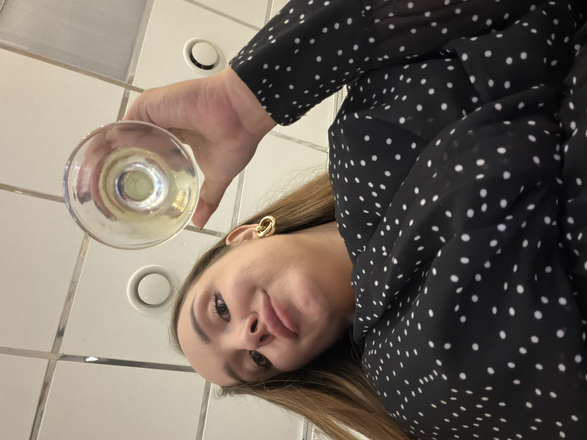 A low-angle, medium shot captures a young woman with long, brown hair holding a clear glass of an amber-colored liquid near her face. She is looking directly into the camera, with a slight smirk. The woman is wearing a black dress with a white polka dot pattern and a gold hoop earring. The background appears to be an indoor space with a white tiled ceiling that has circular vents. The overall lighting is bright, and there are no visible elements that definitively place the location in Chaikovsky, Russia.