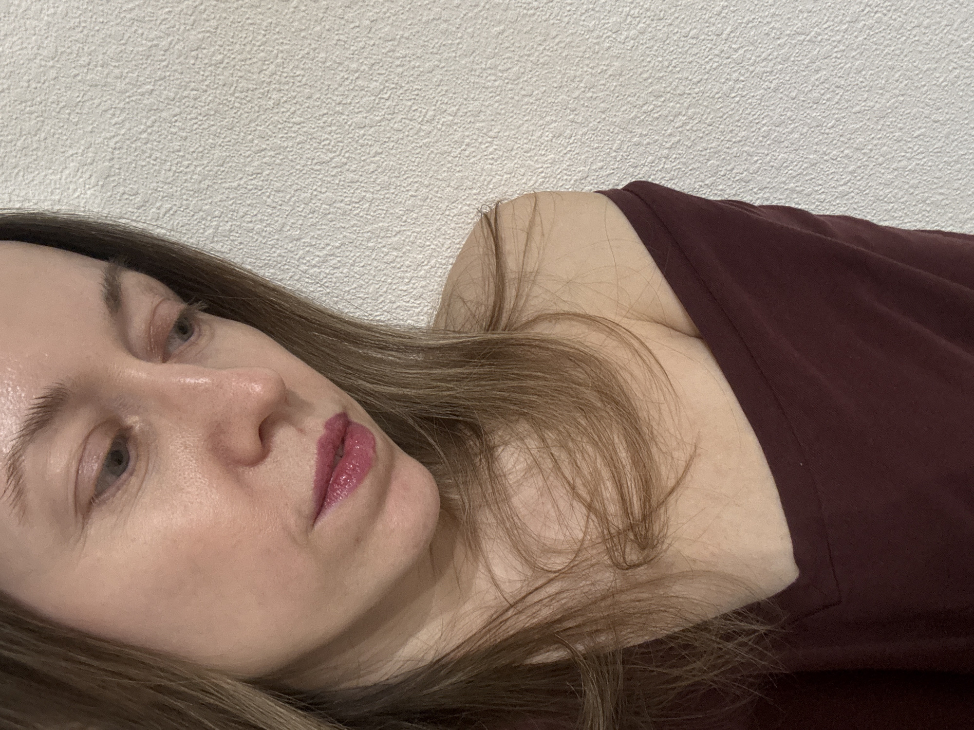 A close-up, high-angle portrait shot depicts the upper body of a light-skinned female individual in Kudrovo, Russia. Her head is tilted downwards and slightly to the left, her gaze directed towards the lower right of the frame. Her long, light brown hair is parted on the left and falls over her right shoulder and chest. Her right eye is partially visible, while her lips are coated with a glossy fuchsia-toned lipstick. She wears a dark maroon V-neck top, which exposes her right shoulder and collarbone. The background consists of a uniformly textured, off-white wall. No interactions or specific actions beyond her pose are observable.